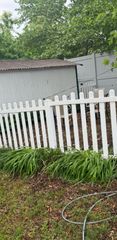 White picket fence with green plants in front of a shed and leafy trees in a backyard.