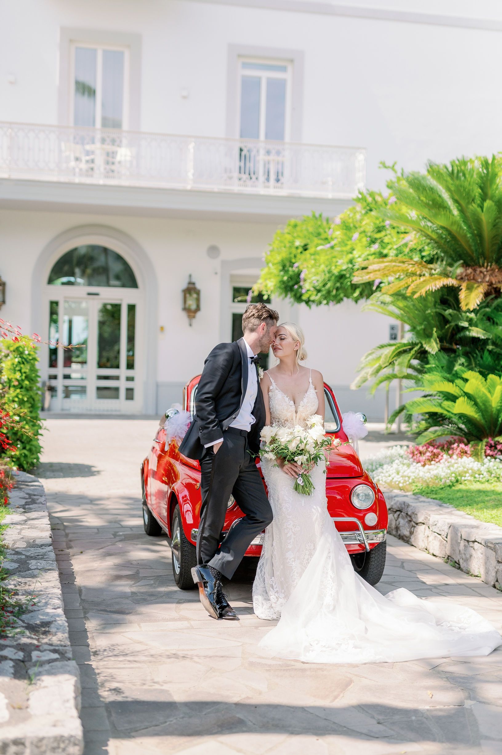 Couple in wedding attire standing by a red vintage car in front of a white house