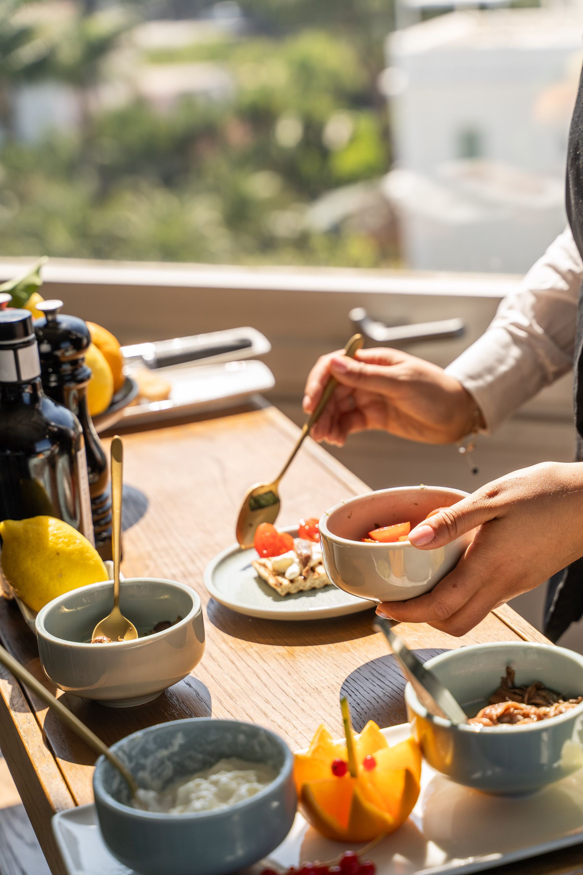 Mani che impiattano un piccolo dessert all'aperto, accanto a ciotole, frutta e condimenti su un tavolo soleggiato.