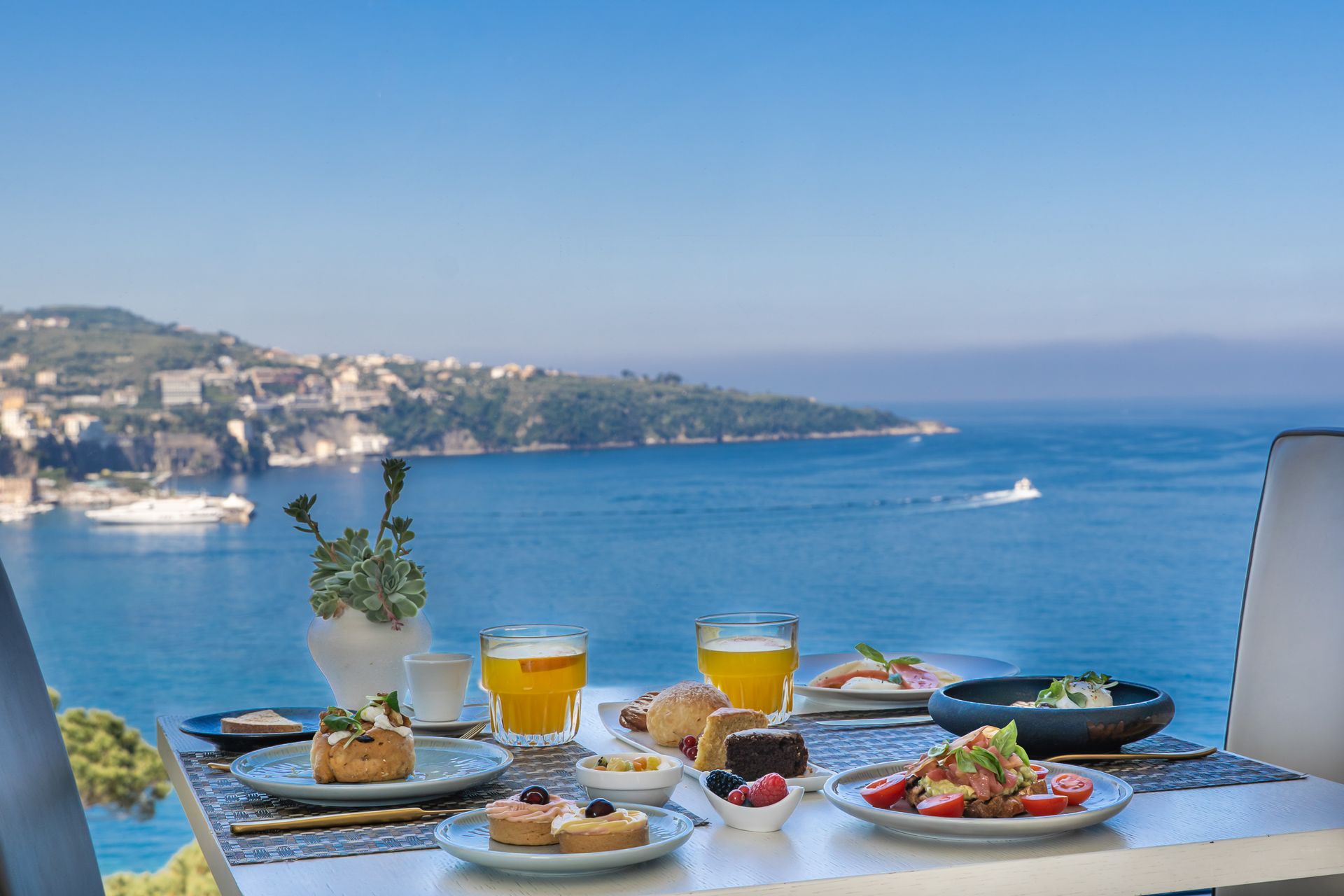 Breakfast on the terrace of Vesuvio Restaurant with coffee, fruit, and pastries overlooking a blue bay and coastal town