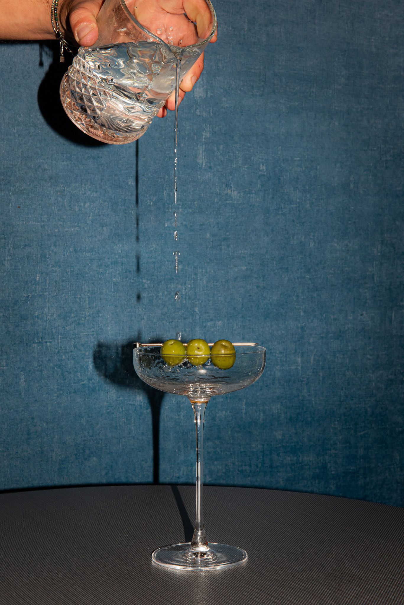 Una mano versa un liquido trasparente da una brocca di vetro in un bicchiere da cocktail con stelo contenente tre olive verdi.