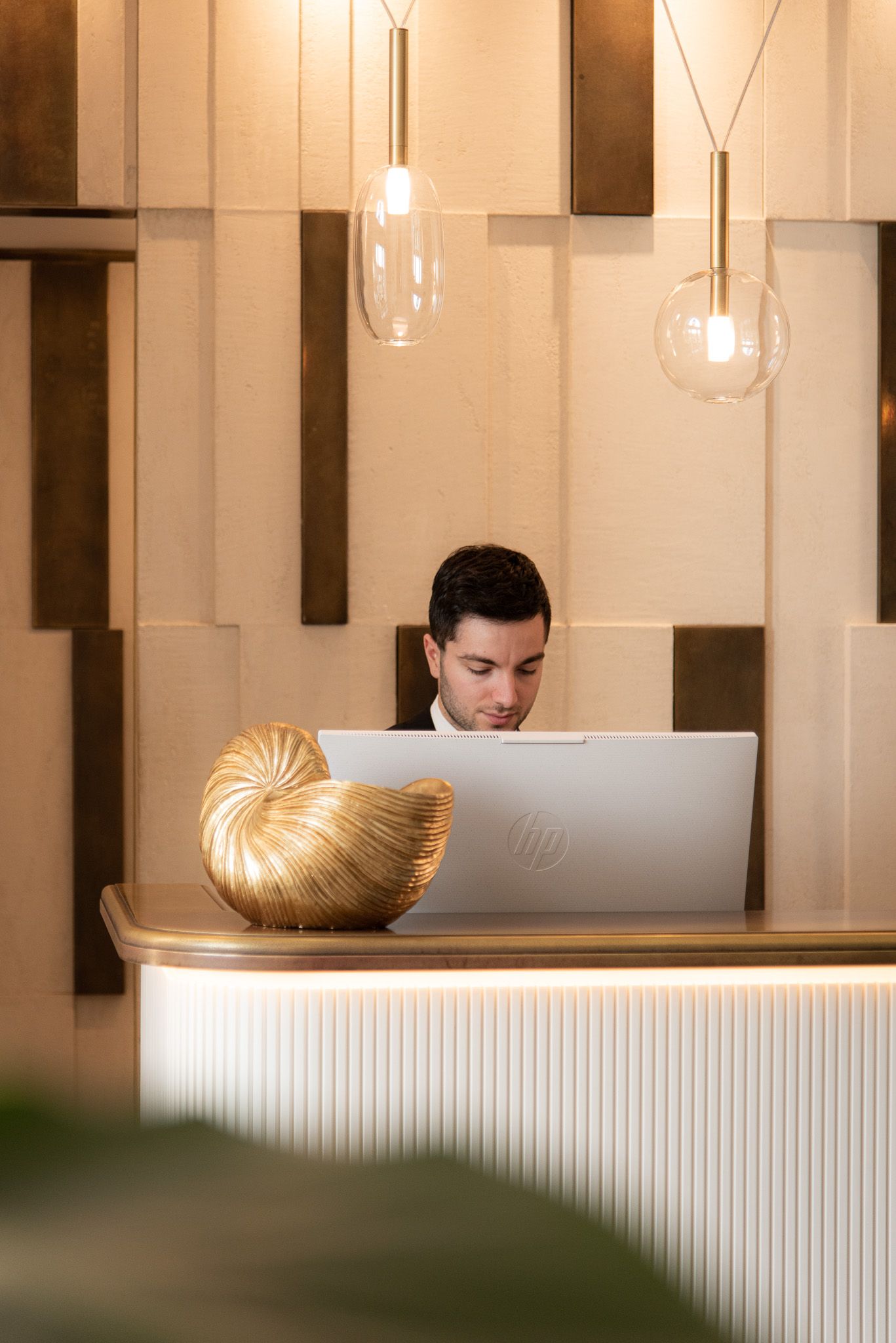 A person working behind a modern, neutral-toned reception desk with a gold decorative object and two hanging pendant lights.