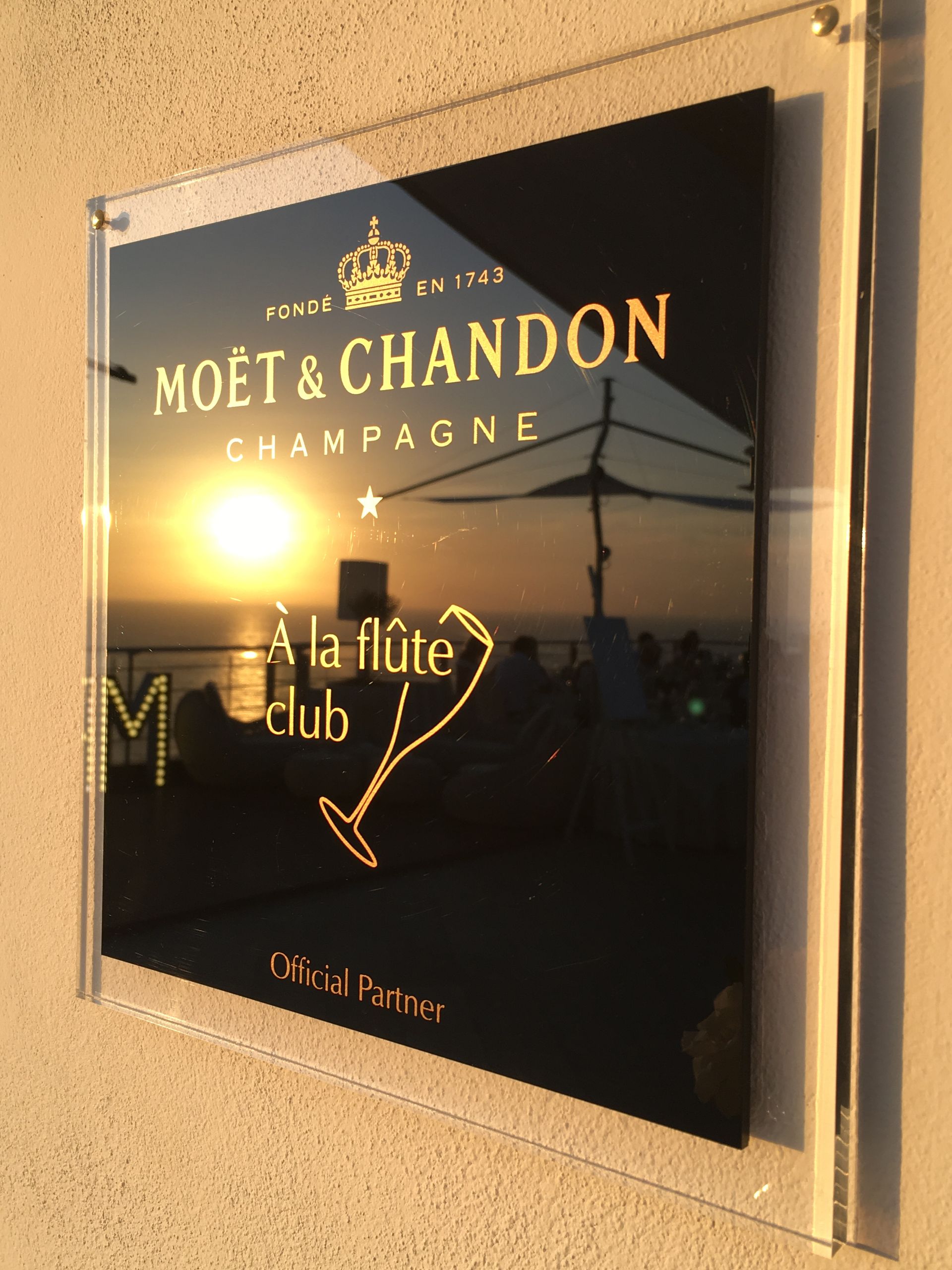 Un'insegna a parete per Moët & Chandon, con testo dorato su acrilico nero che riflette un tramonto sul mare.