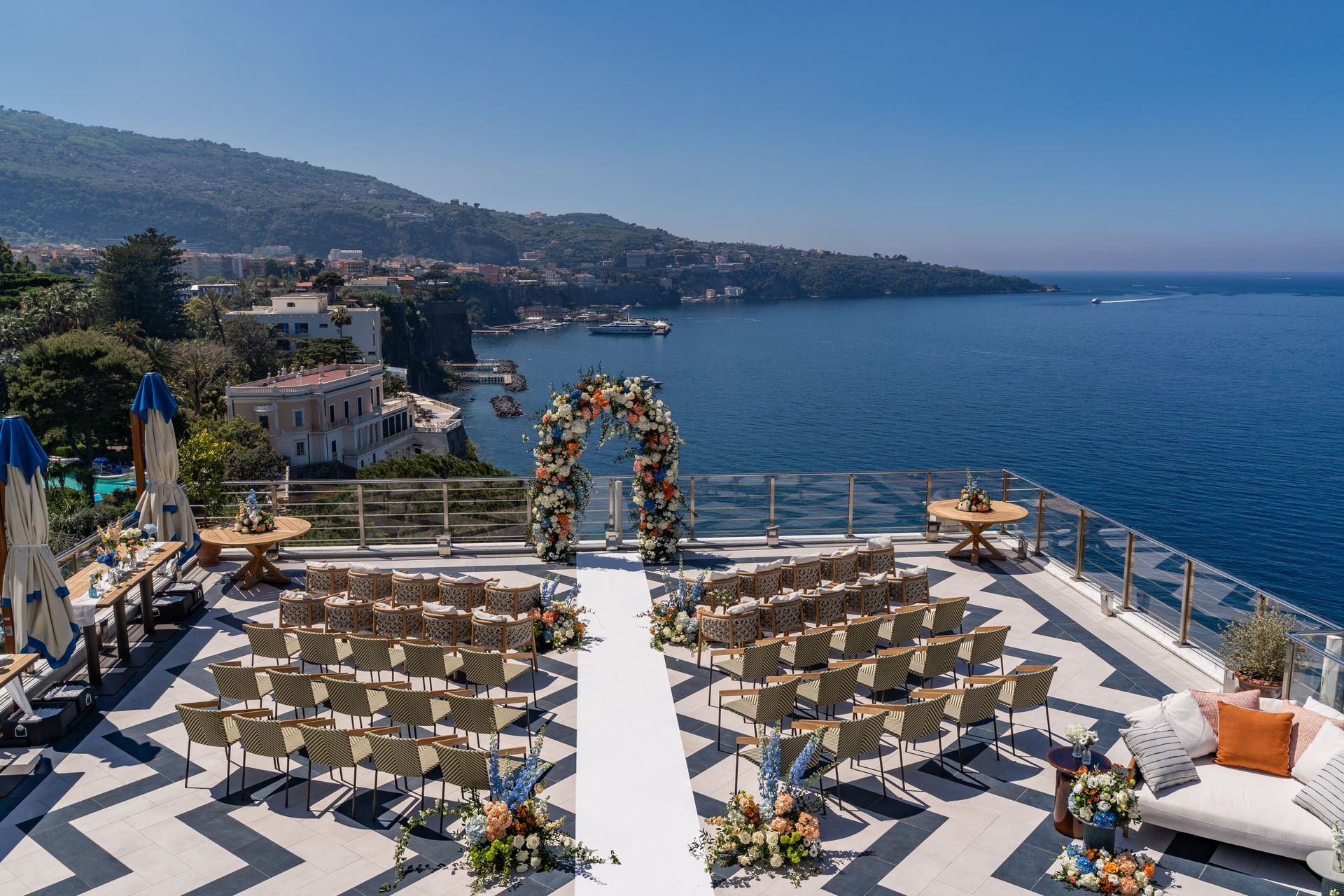 A sunset view of a restaurant patio overlooking the ocean with guests seated at tables and lounging on white sofas.