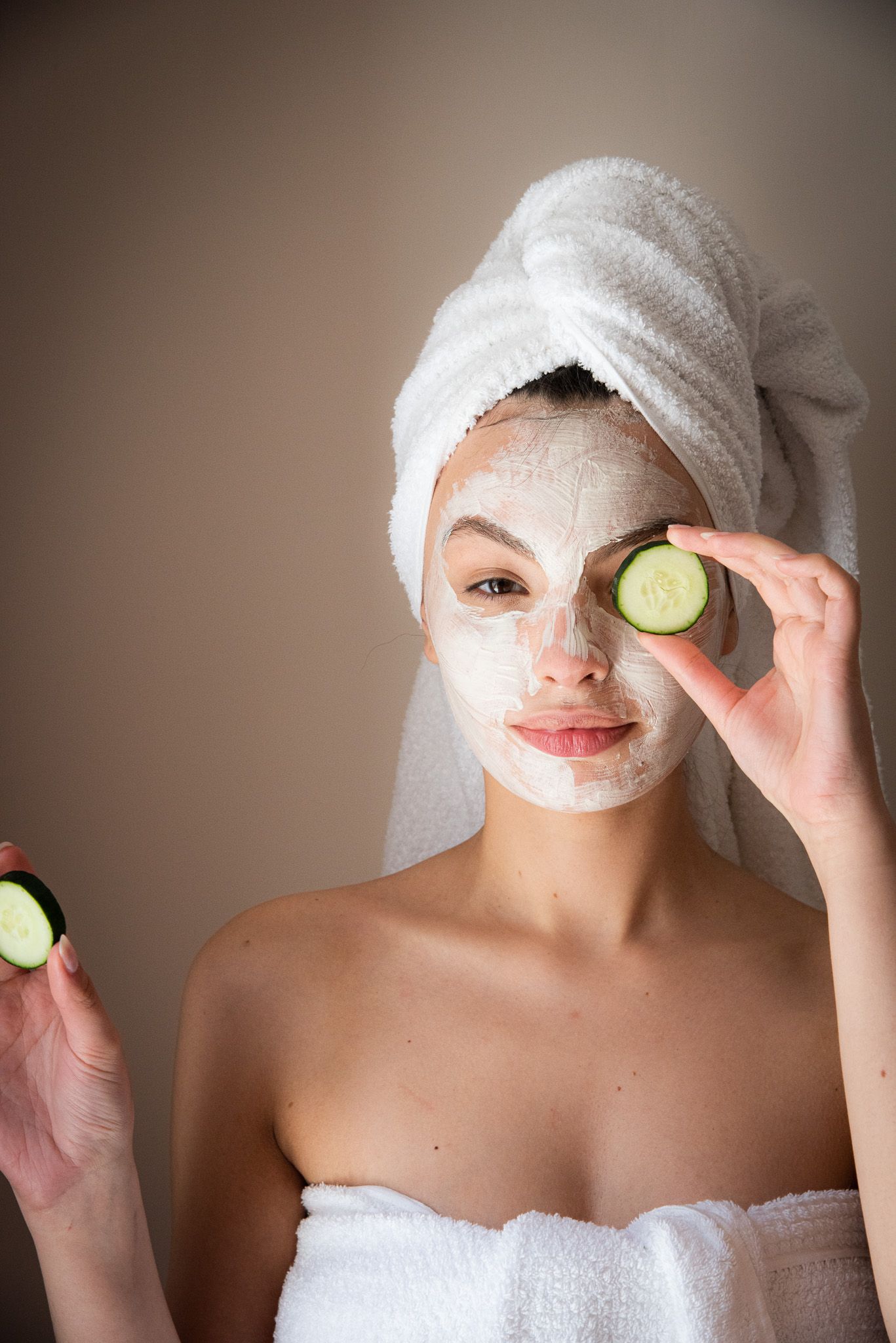 In un ambiente che ricorda una spa, una persona avvolta in un asciugamano bianco e con una maschera per il viso si copre gli occhi con delle fette di cetriolo.