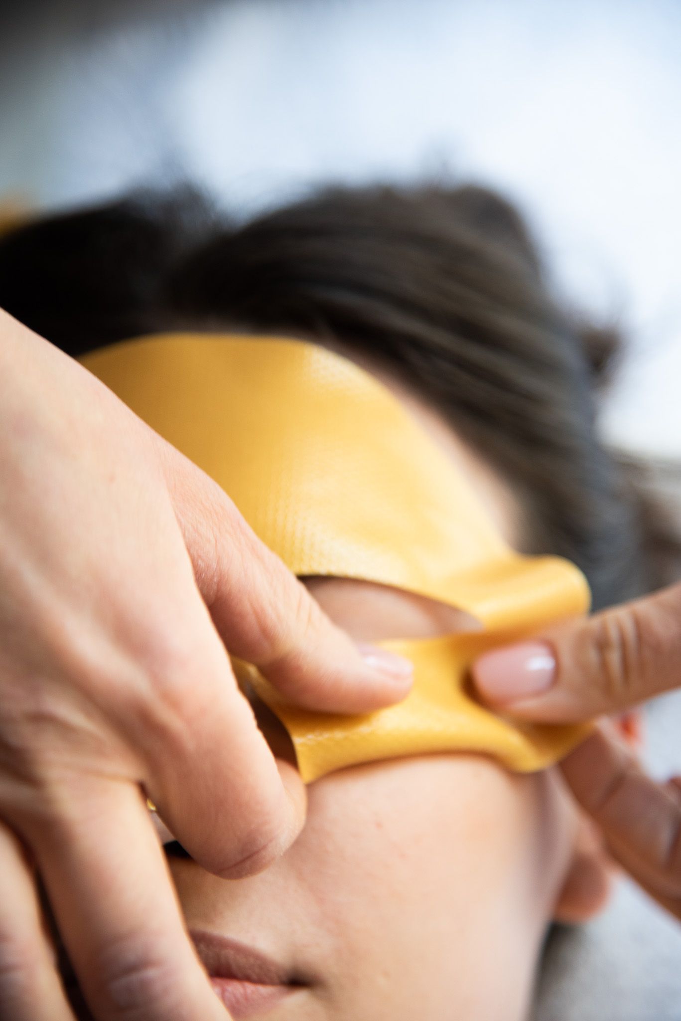 Un paio di mani applicano con cura una maschera per il viso liscia, di colore giallo dorato, sulla parte superiore del volto di una persona.