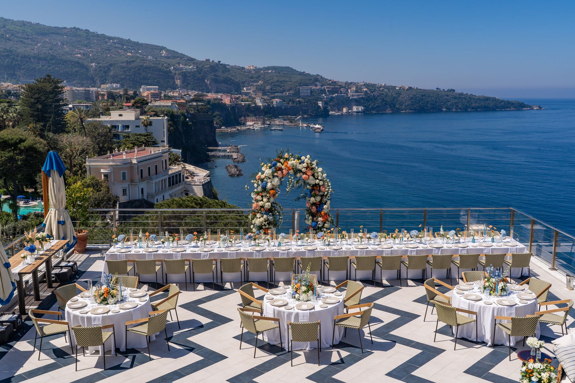 Elegant seaside terrace set for a wedding reception, overlooking a blue bay and coastal hillside.