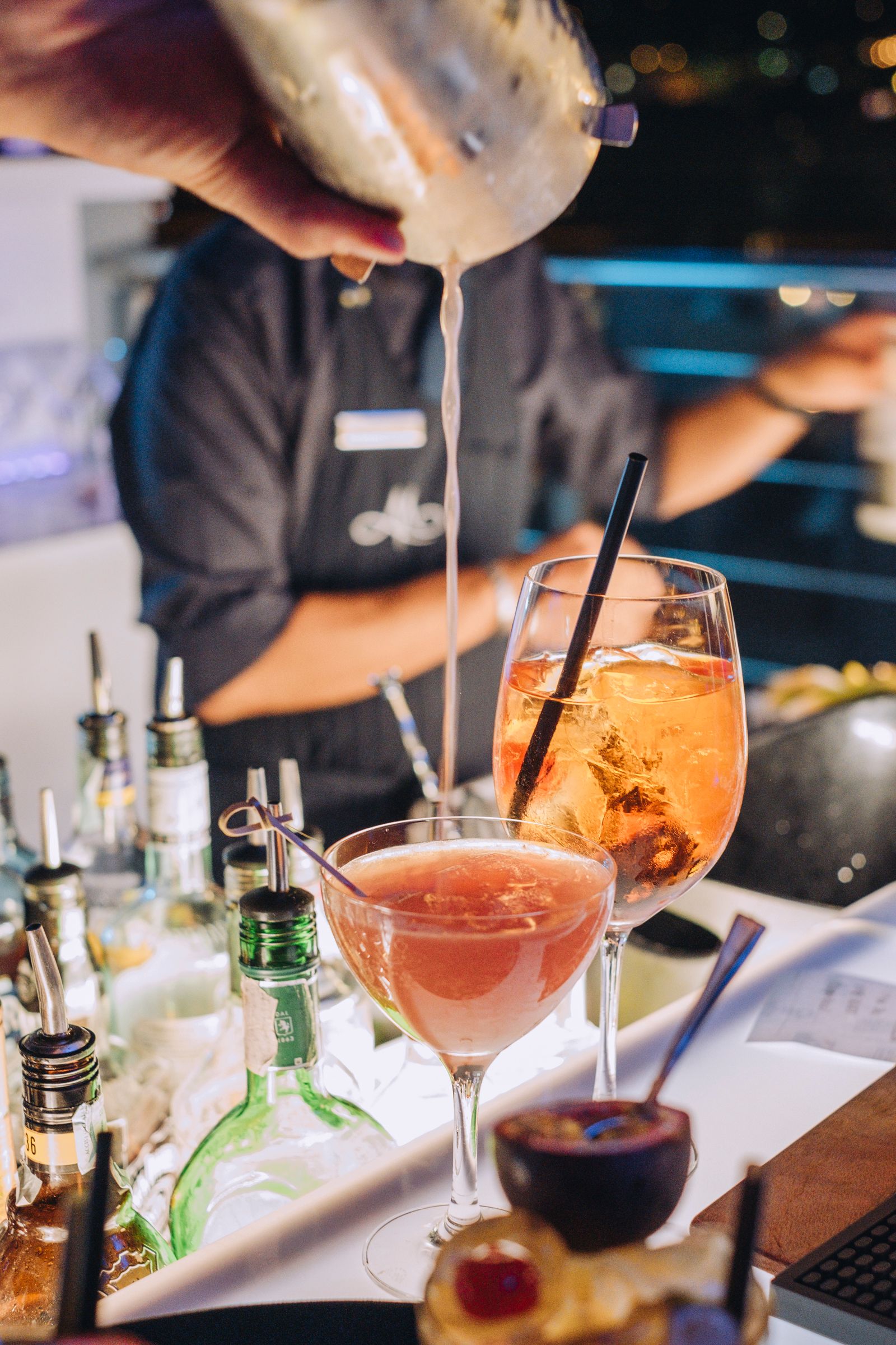 In un bar affollato e scarsamente illuminato, dove sono già presenti bevande già pronte, un barista versa un cocktail da uno shaker di metallo in un bicchiere.