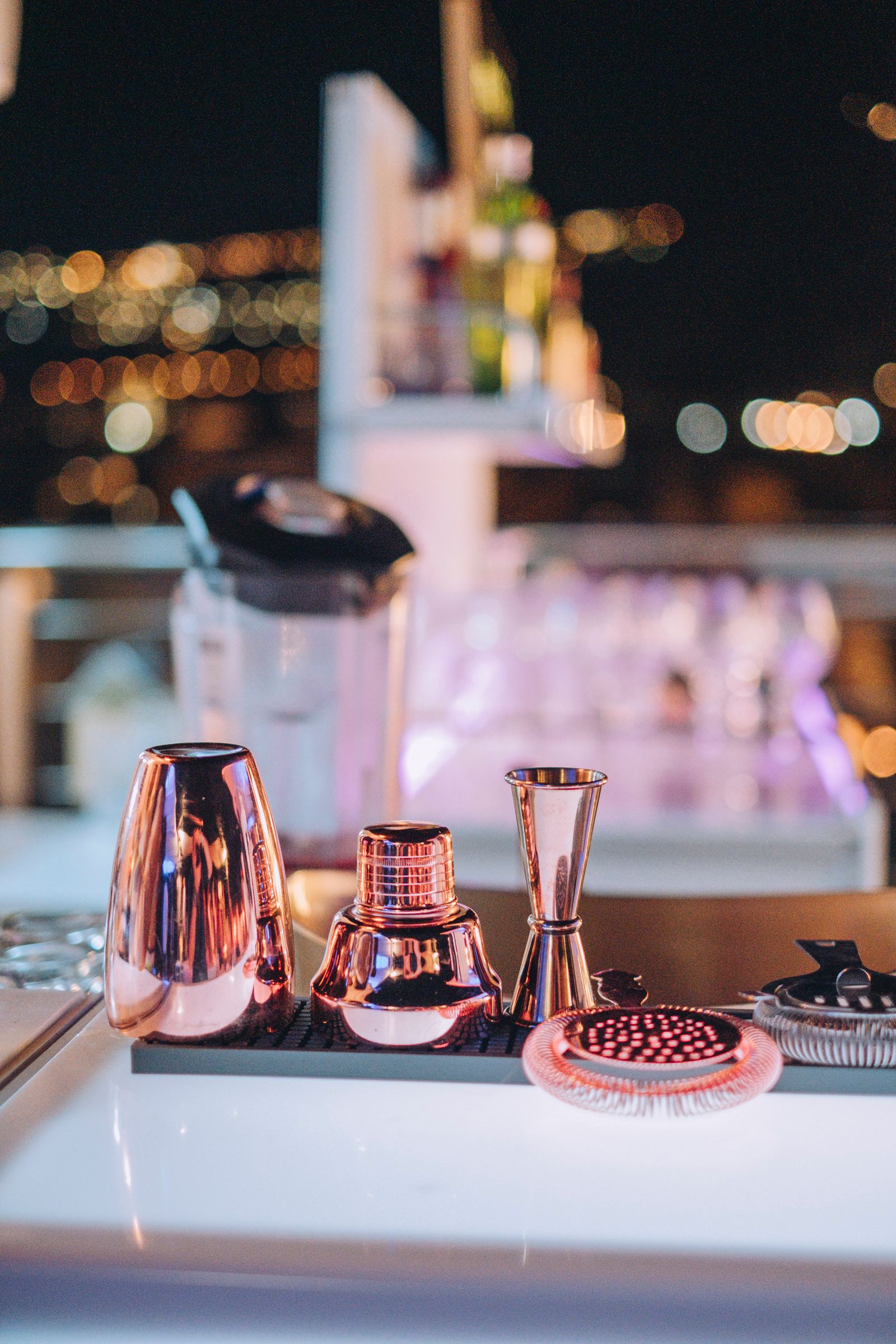 Utensili da cocktail in rame su un vassoio luminoso, sullo sfondo di uno skyline serale sfocato, illuminato da calde luci bokeh.