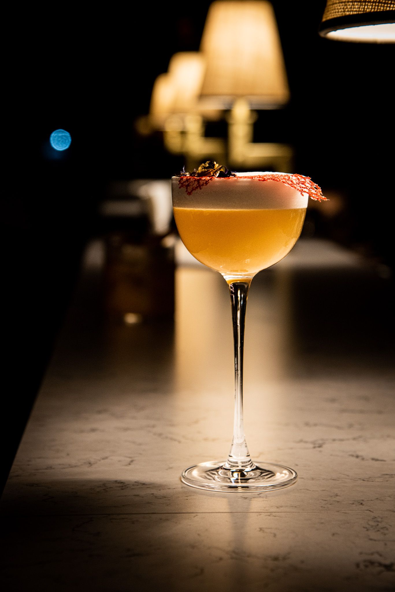 Un cocktail dorato con una spuma in superficie, guarnito con petali secchi, è appoggiato sul bancone di un bar, illuminato da luci soffuse e calde.