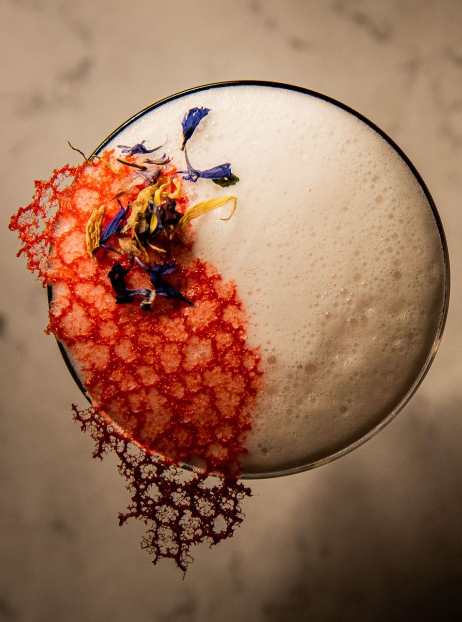 Vista dall'alto di un cocktail sormontato da una spuma, guarnito con una cialda di pizzo rosso acceso e petali di fiori commestibili sparsi.