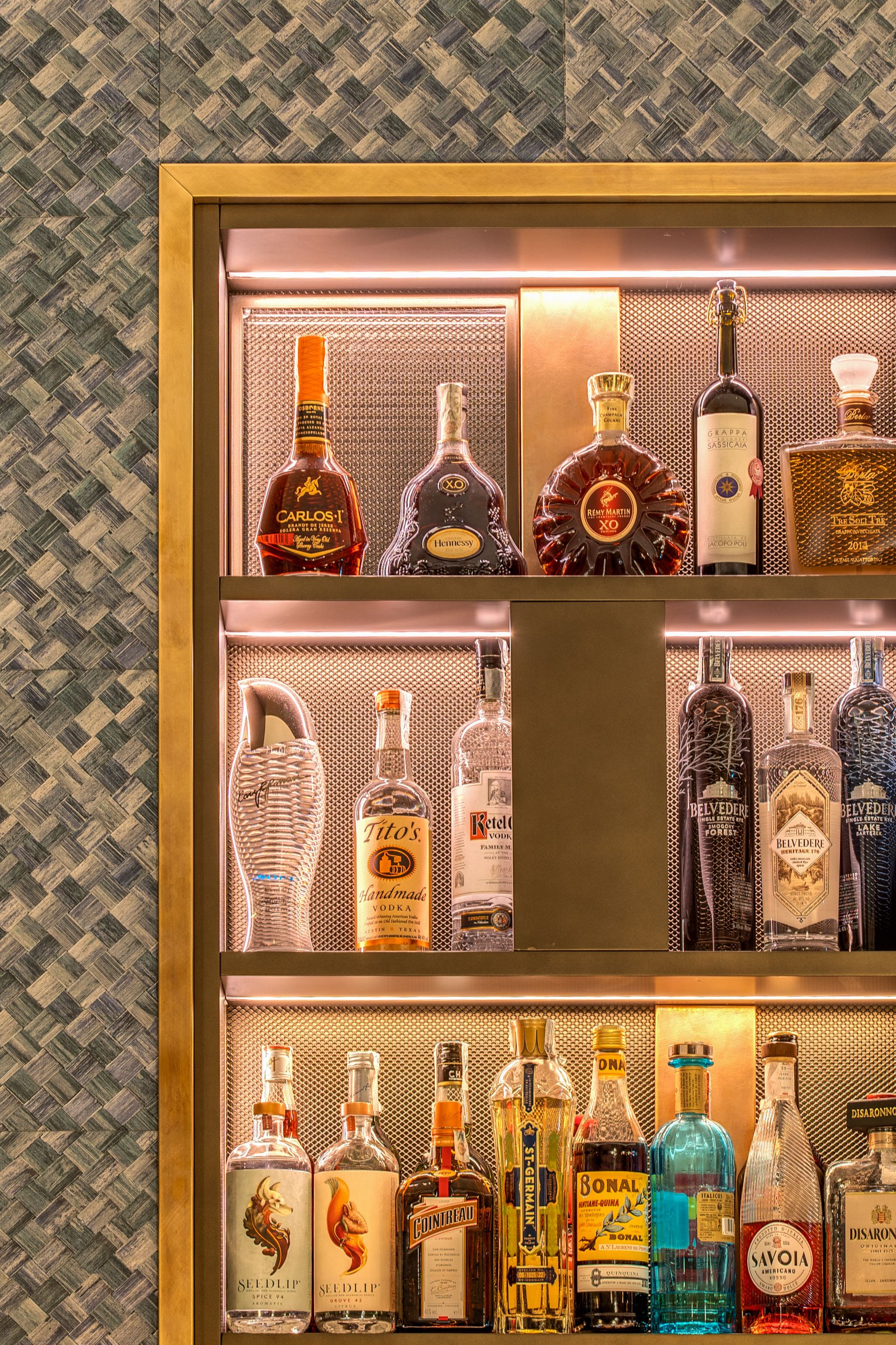 Uno scaffale da bar retroilluminato con tre livelli di bottiglie di liquori assortite, su uno sfondo con texture e motivo metallico.