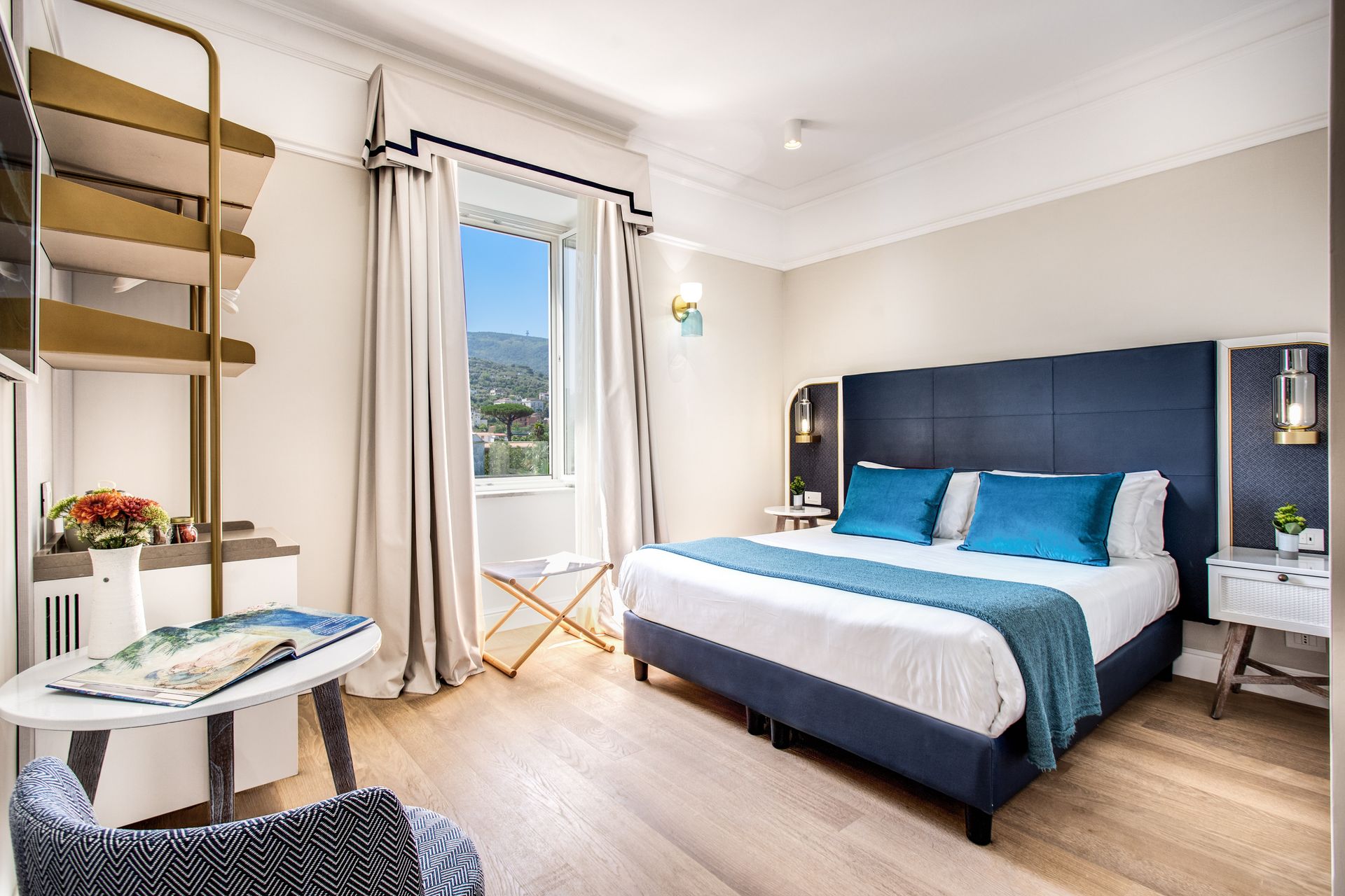Una camera da letto con un letto caratterizzato da una testiera blu scuro, lenzuola bianche, cuscini color verde acqua, una finestra con tende e un comodino.