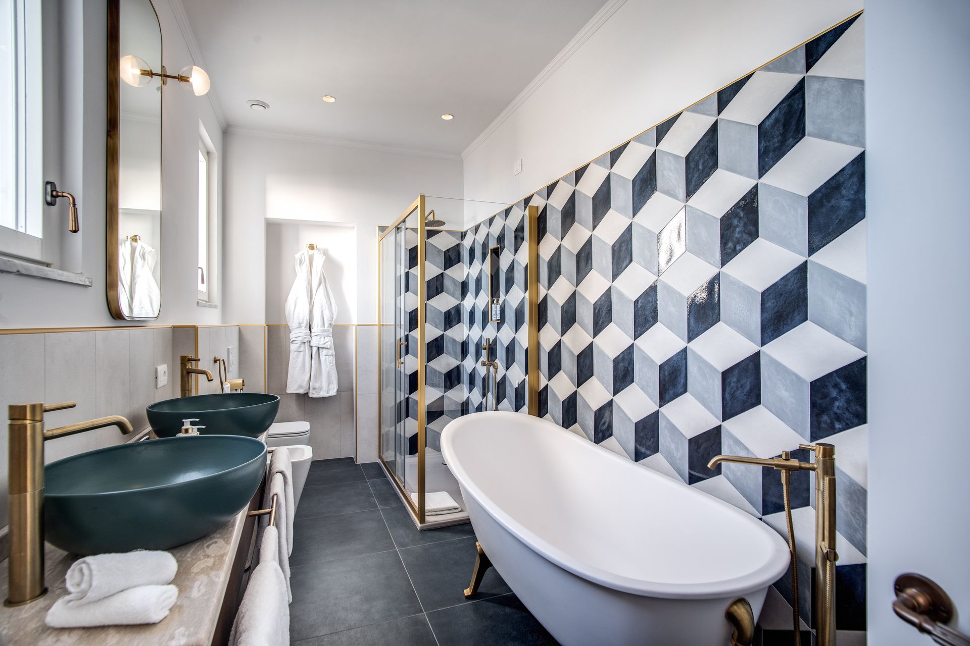 Un bagno moderno con vasca con piedini, lavabi verde scuro, rubinetteria dorata e una parete rivestita con piastrelle geometriche blu e bianche.
