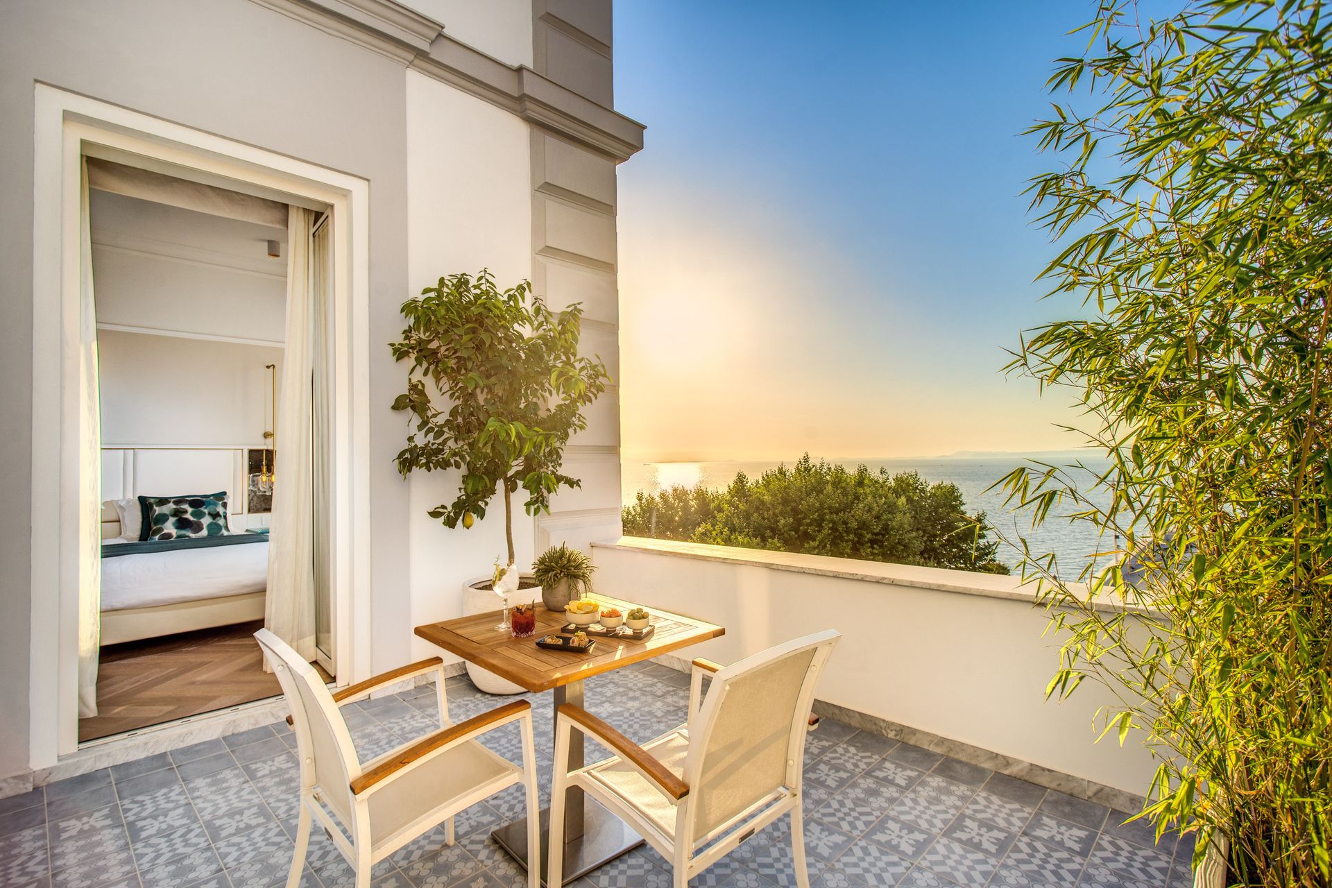 Un balcone soleggiato con due sedie e un tavolino di legno, affacciato sull'oceano al tramonto, vicino a una porta della camera da letto aperta.
