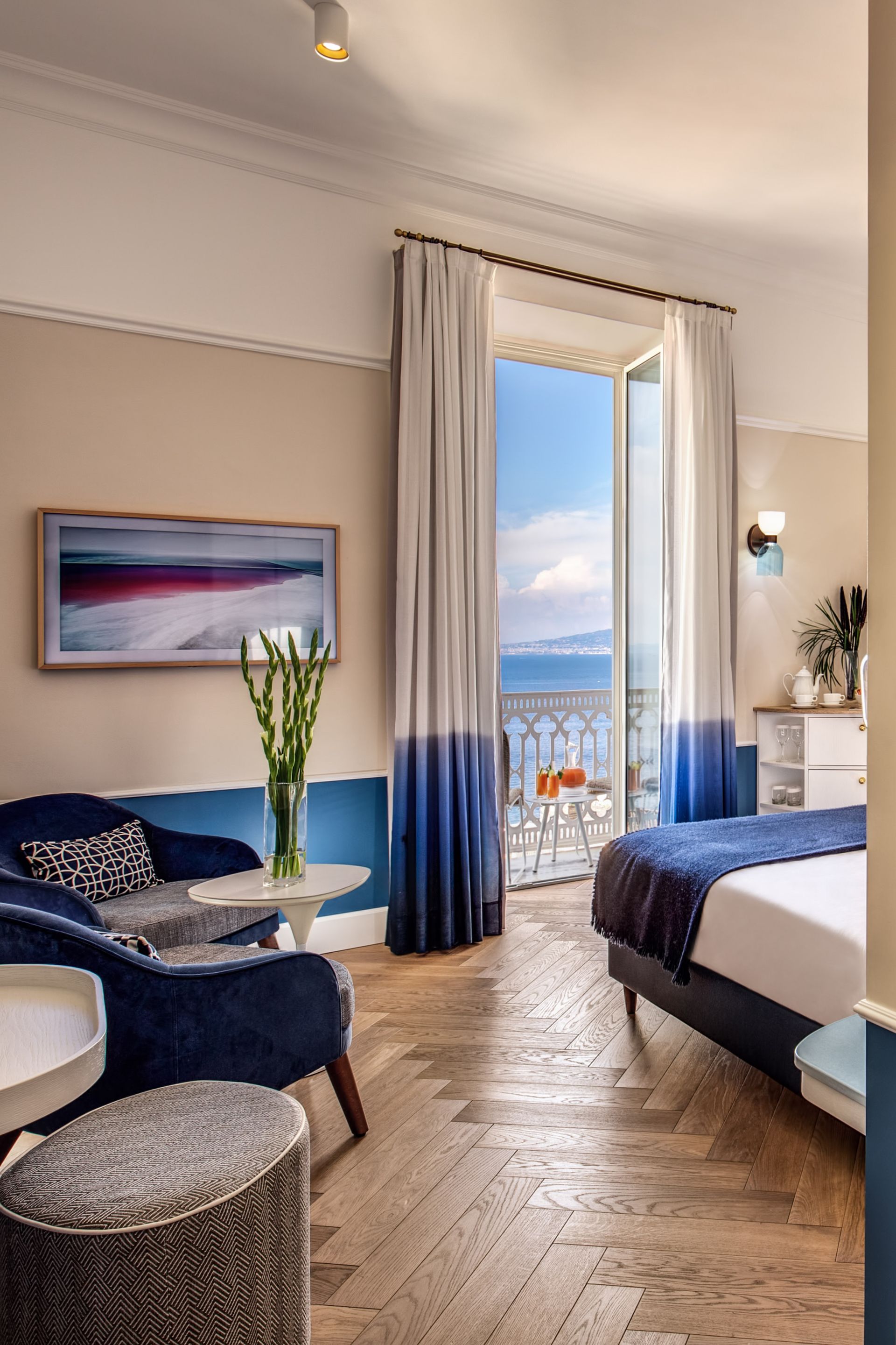 Una camera d'albergo con pavimento in legno a spina di pesce, un divano in velluto blu, un letto e porte finestre che si aprono sul balcone con vista sul mare.