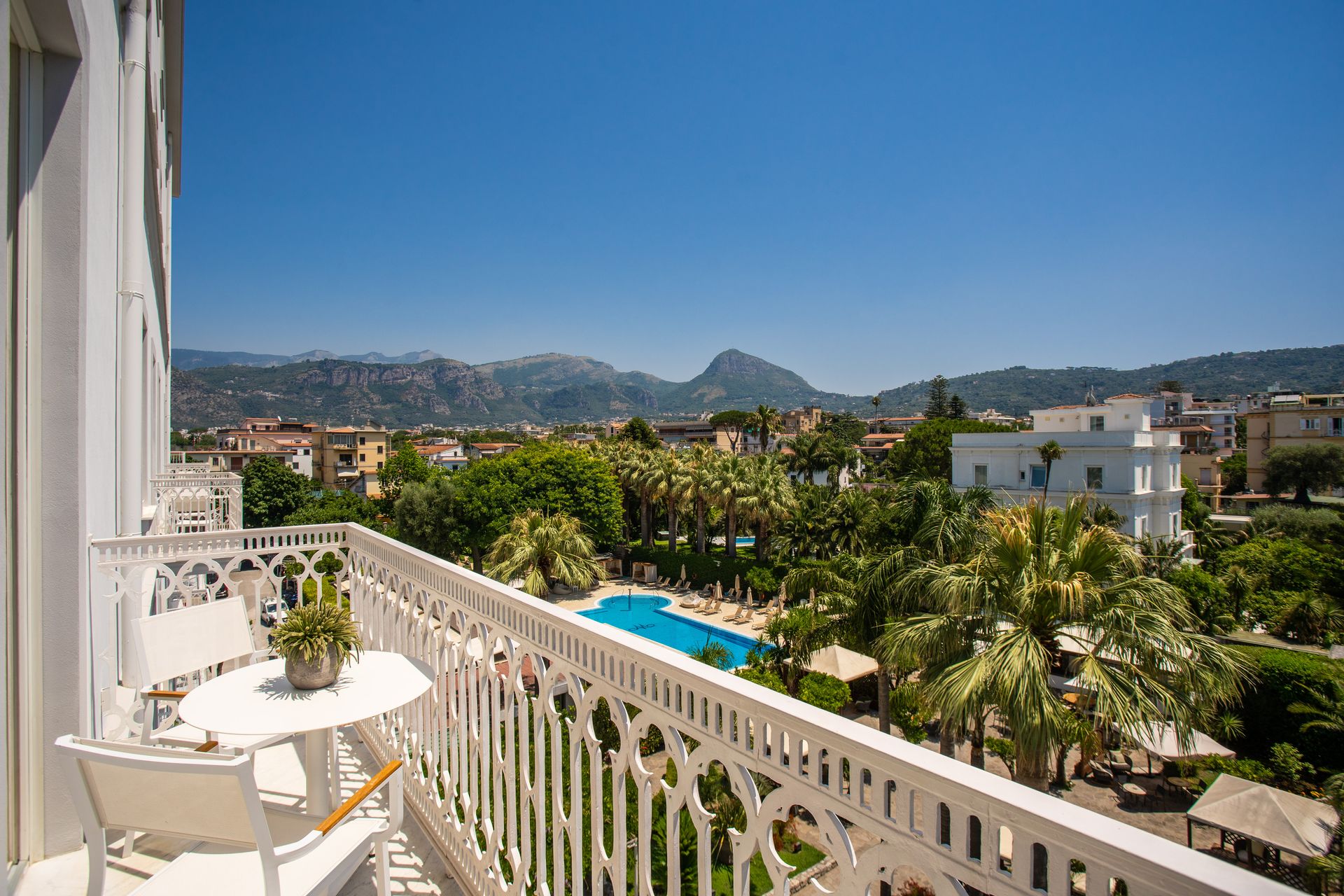 Un balcone bianco con tavolo e sedie che si affaccia su una piscina, palme e una catena montuosa sotto un cielo limpido.