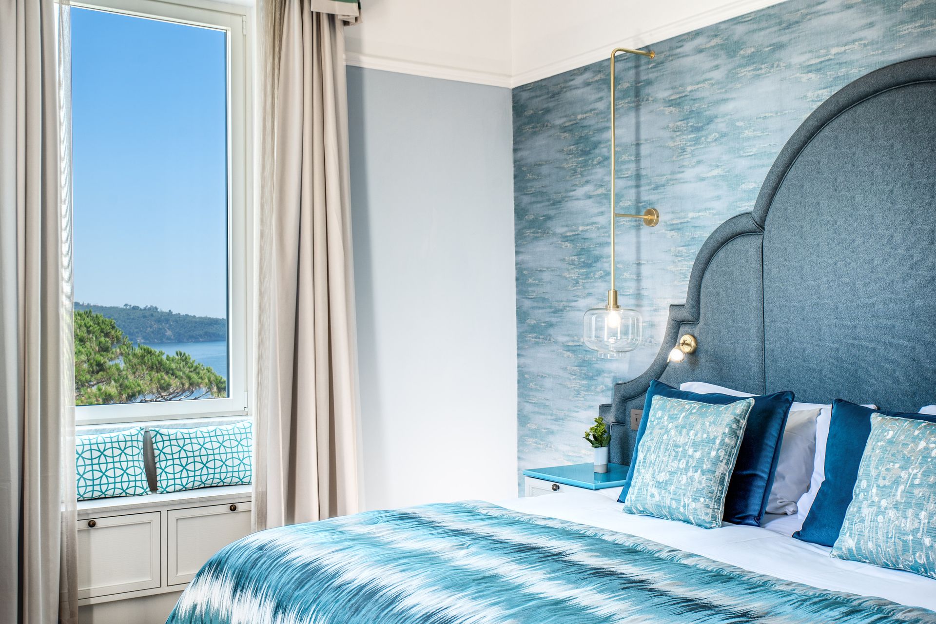 Una camera da letto a tema blu con una splendida vista sull'oceano attraverso una finestra, caratterizzata da biancheria da letto a fantasia e una grande testiera.