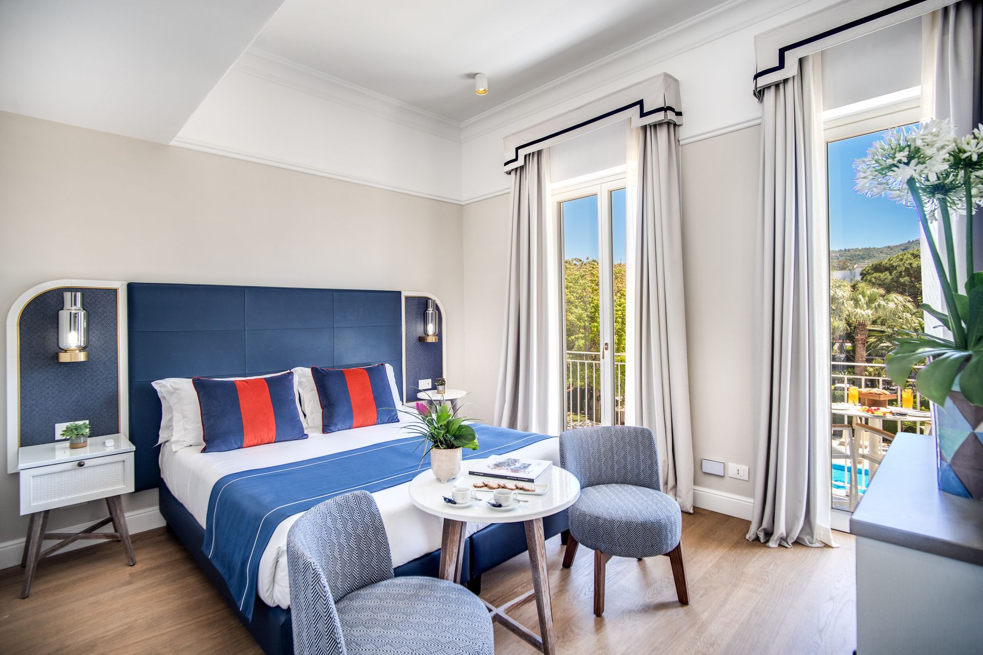 Una luminosa camera d'albergo con un letto blu, due sedie decorate, un tavolino e porte finestre che si aprono su un balcone.