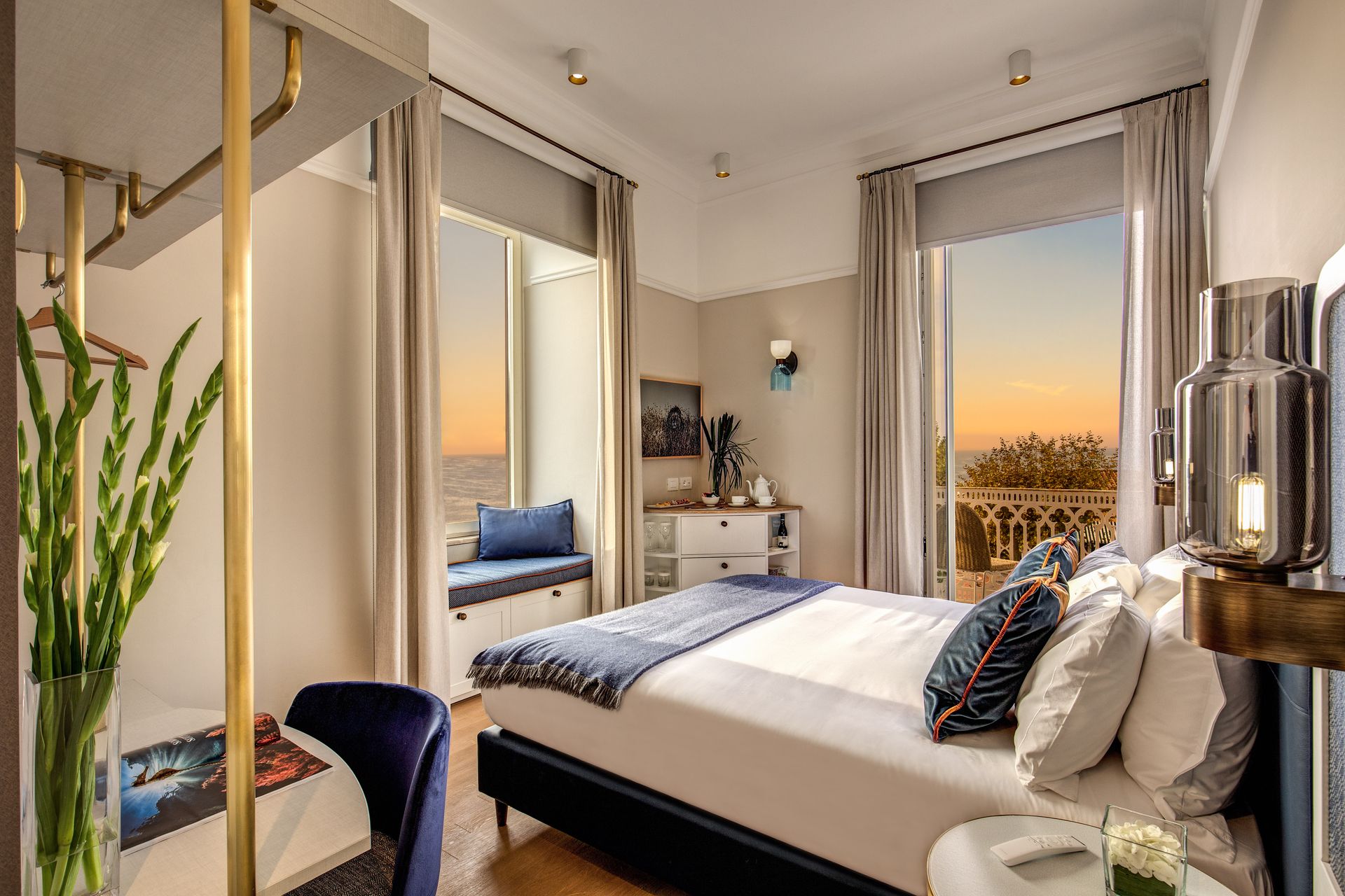 Una moderna camera d'albergo con un letto lussuoso, un angolo salotto accanto alla finestra, dettagli dorati e vista sul tramonto.