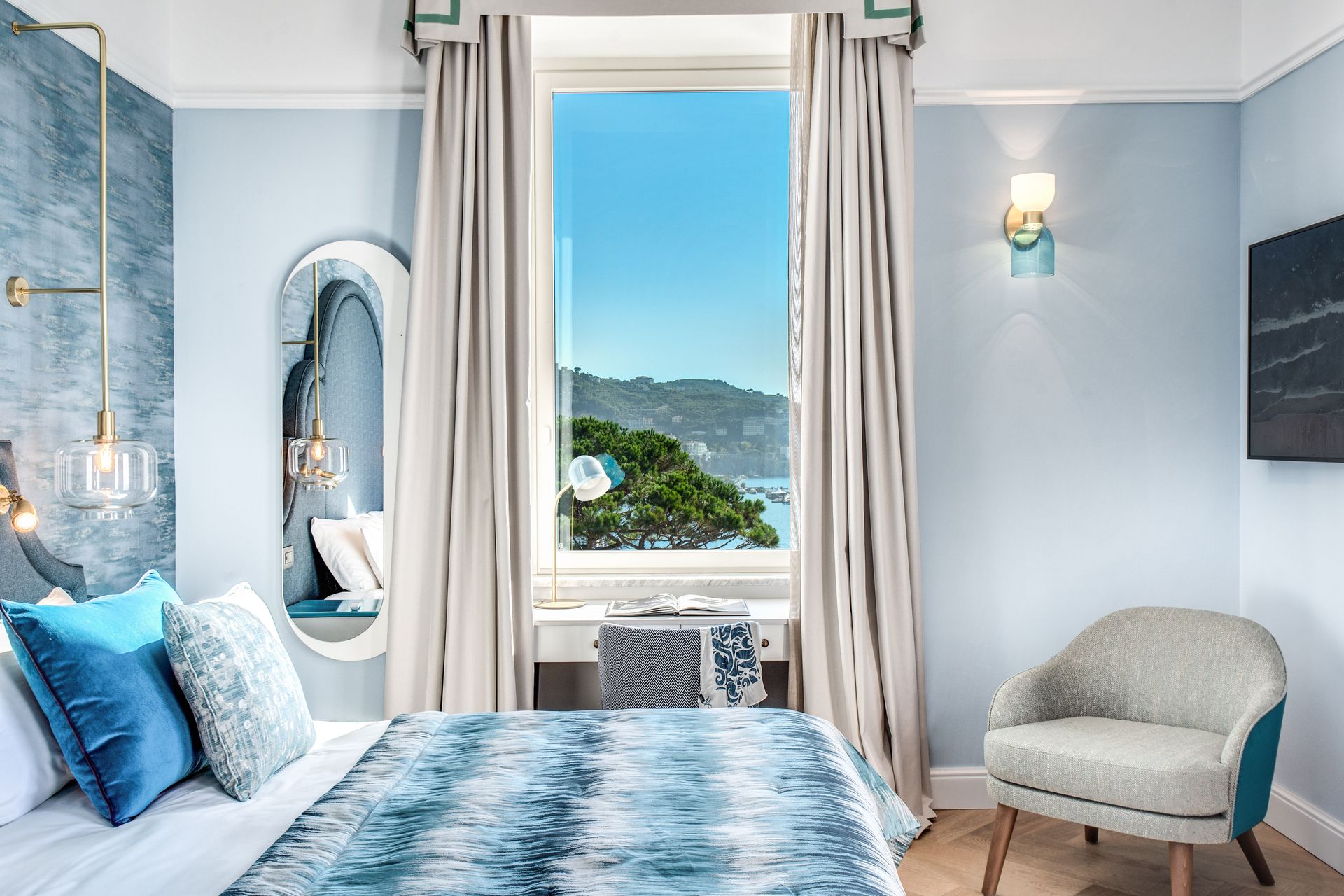 Una luminosa camera d'albergo con un copriletto a fantasia blu, uno specchio sul comodino e una finestra con vista su una verde collina e sull'oceano.