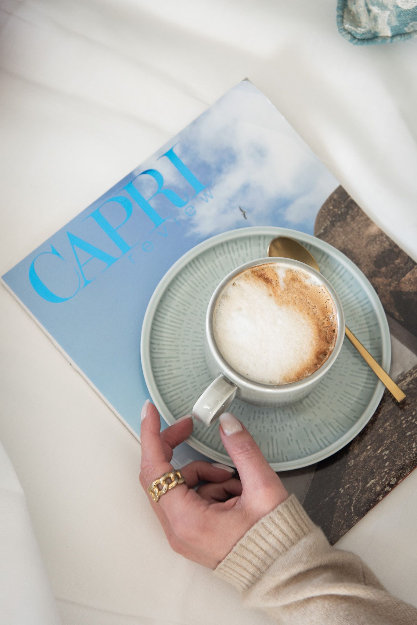 Una mano con un anello d'oro regge una tazza di cappuccino in ceramica su un piattino dalla superficie ruvida, sopra una rivista intitolata 