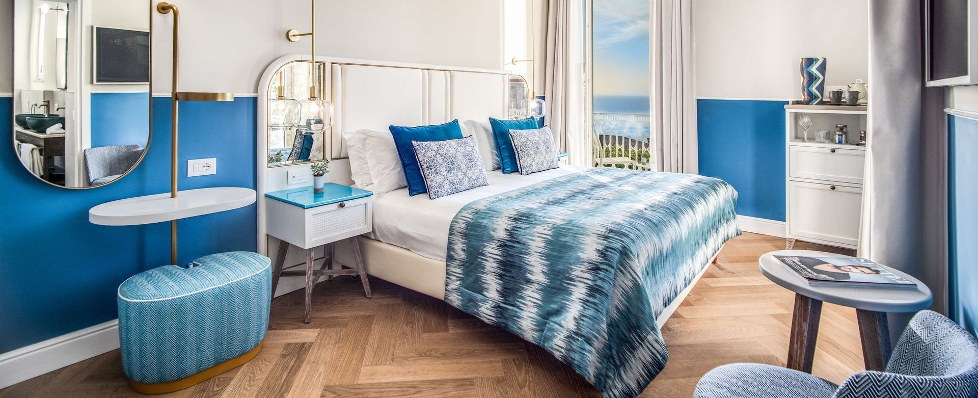 A bright, stylish bedroom with blue walls, a patterned blue bedspread, wooden floors, and a view of the sea from the window.