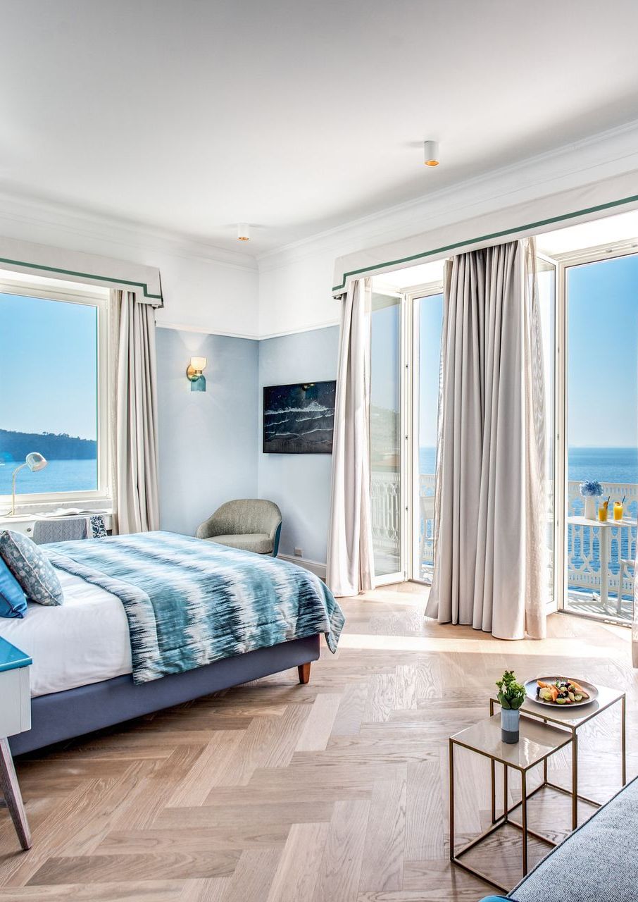 Una camera da letto luminosa con un letto a fantasia blu, pareti azzurre, pavimento in legno a spina di pesce e vetrate a tutta altezza con vista sull'oceano.