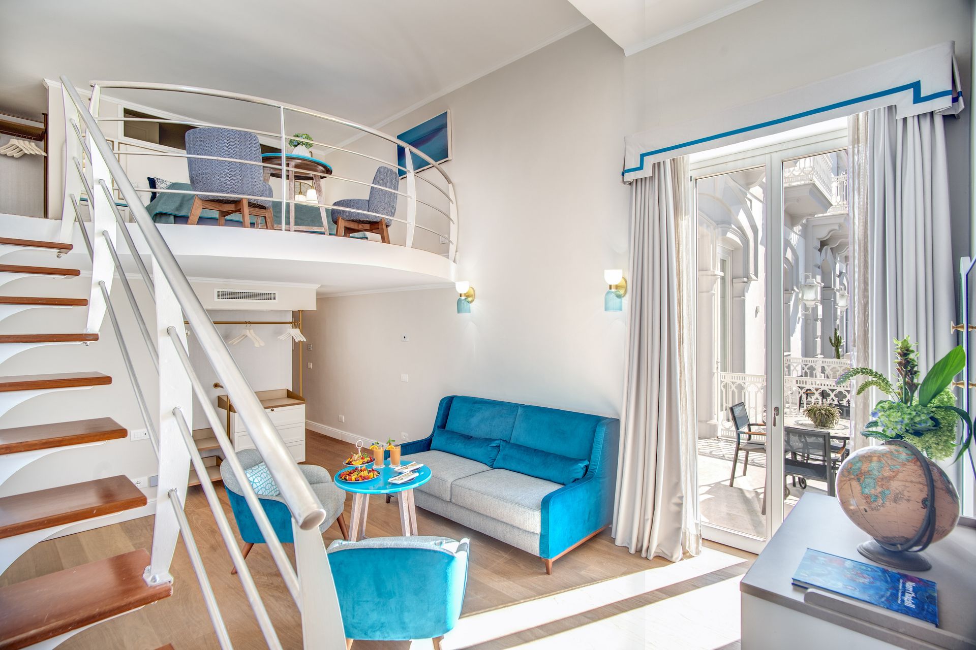Un luminoso loft con divano blu, poltrone, una scala in legno e un balcone visibile attraverso porte a vetri.