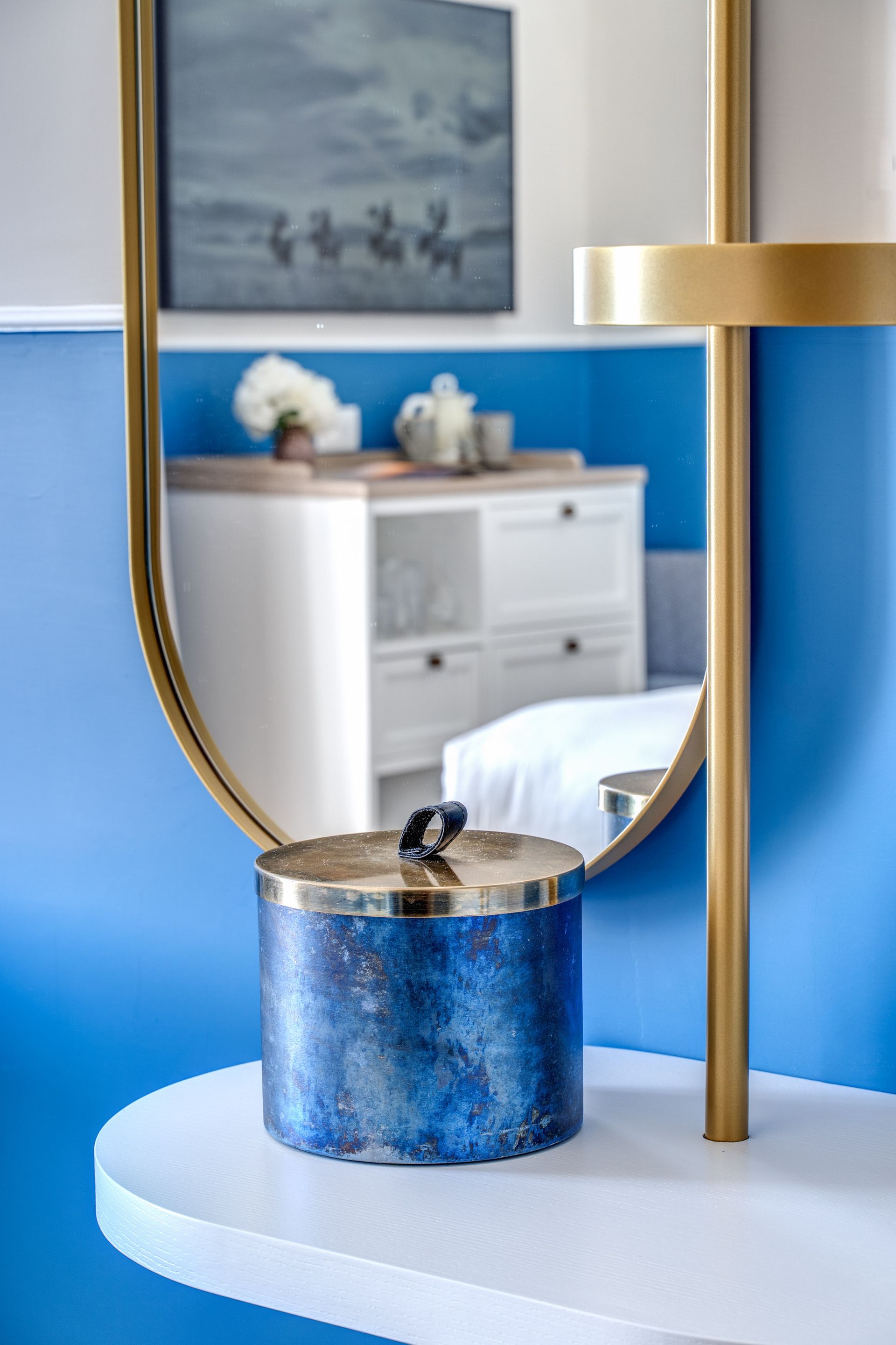 Un contenitore blu con coperchio metallico è appoggiato su una mensola bianca sospesa di fronte a uno specchio rotondo con cornice dorata.