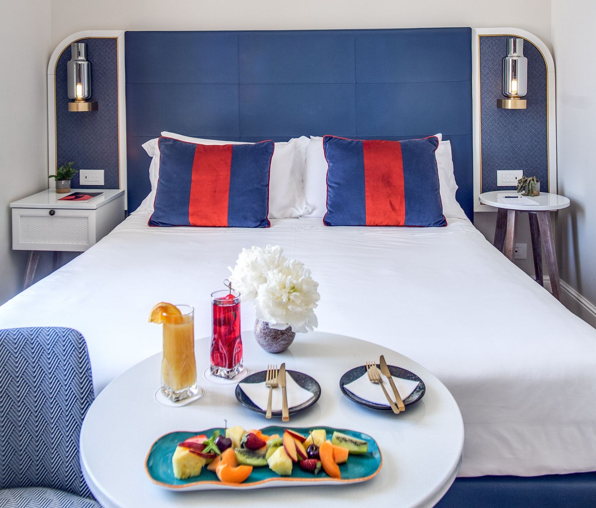 Una camera d'albergo è arredata con un letto con testiera blu, lenzuola bianche e cuscini a righe rosse, mentre sul tavolino si trovano frutta e bevande.