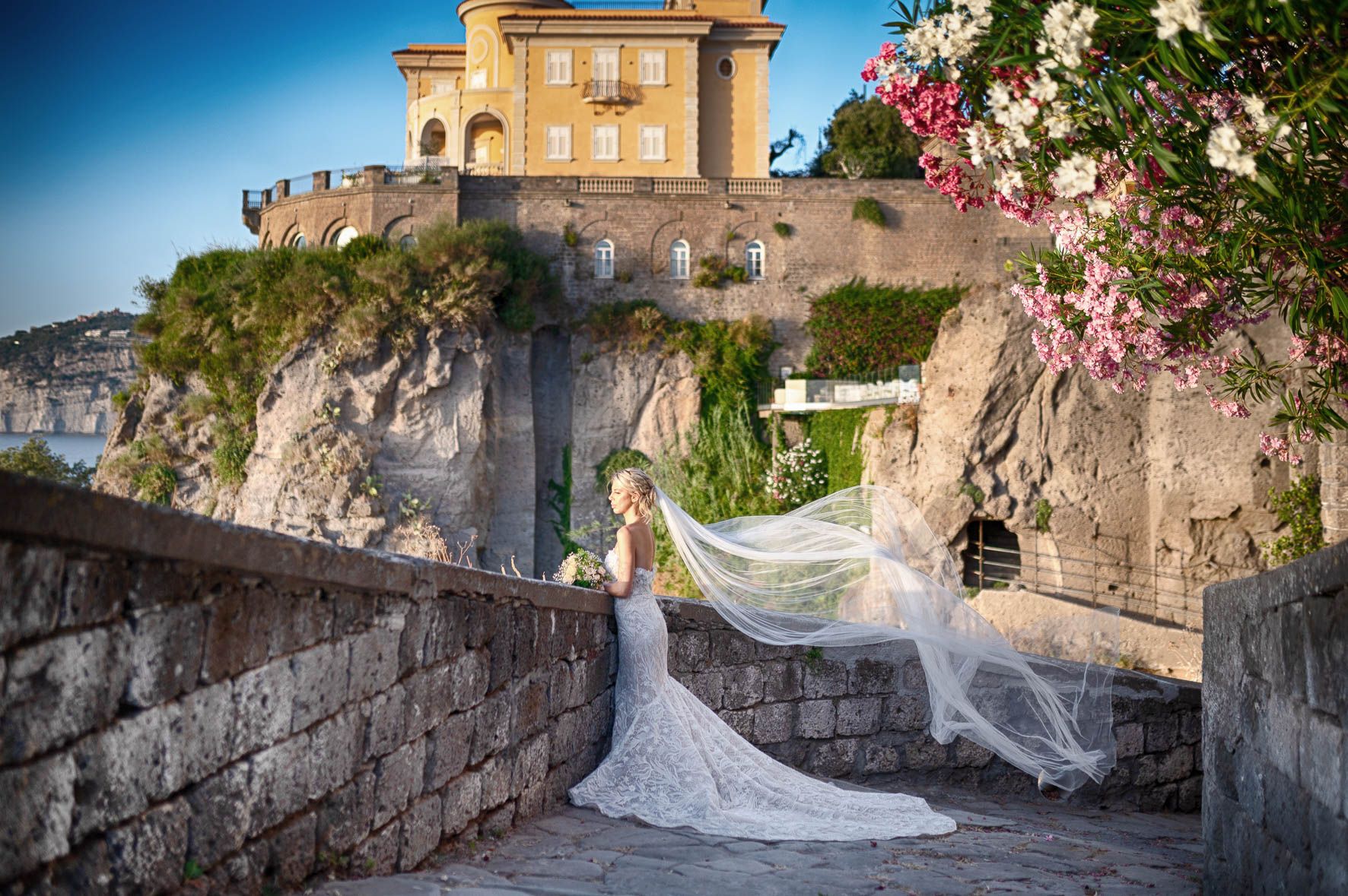 Sposa in abito bianco e velo su un sentiero di pietra accanto a una villa a picco sul mare, con fiori rosa