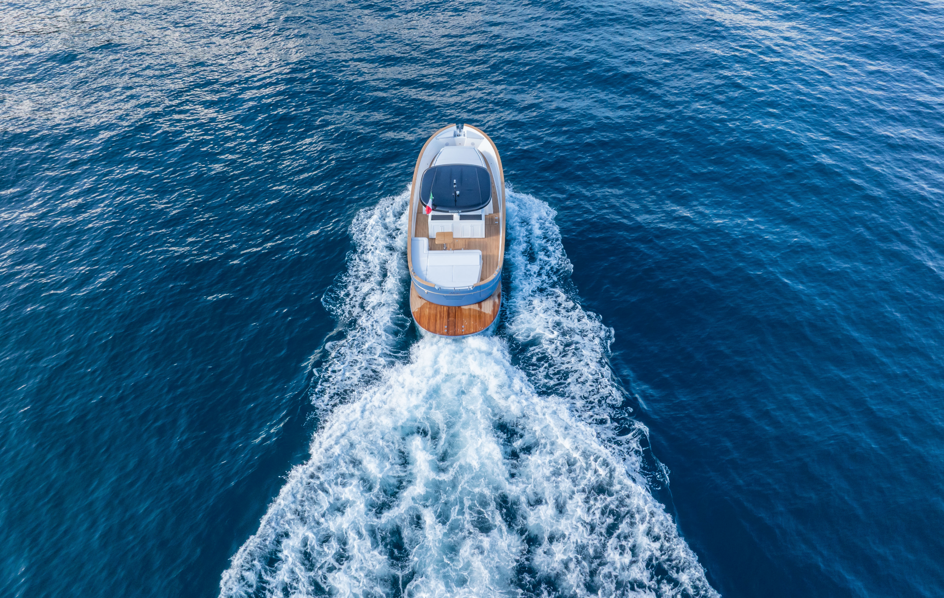 Una veduta aerea di un motoscafo che solca le acque blu intenso, lasciandosi alle spalle un'ampia scia bianca.