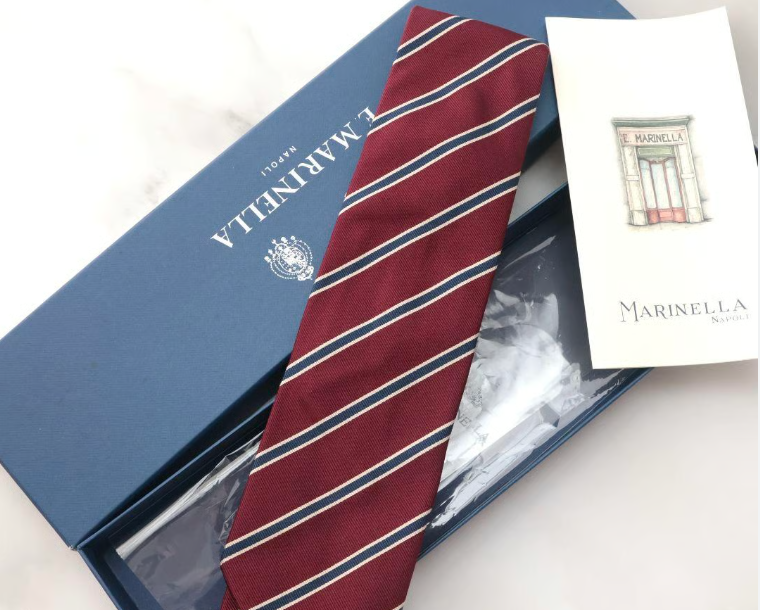 Una cravatta a righe bordeaux è riposta nella sua confezione regalo blu navy di E. Marinella Napoli, con un biglietto informativo del marchio.