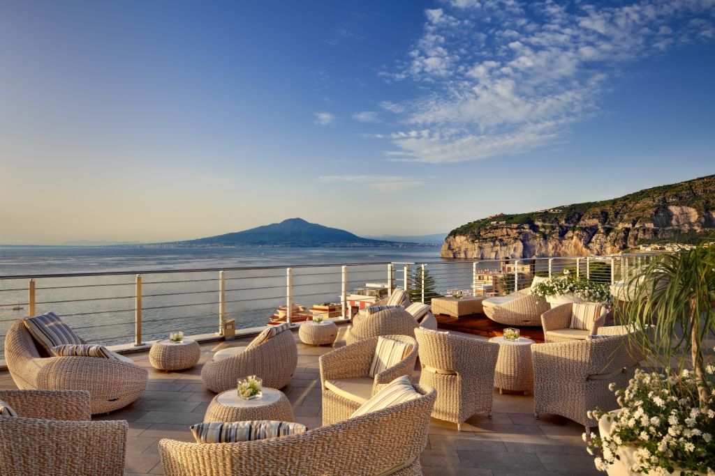 Una lussuosa terrazza con vista sull'oceano e sulle scogliere, arredata con lettini prendisole in rattan sotto un cielo azzurro e limpido.