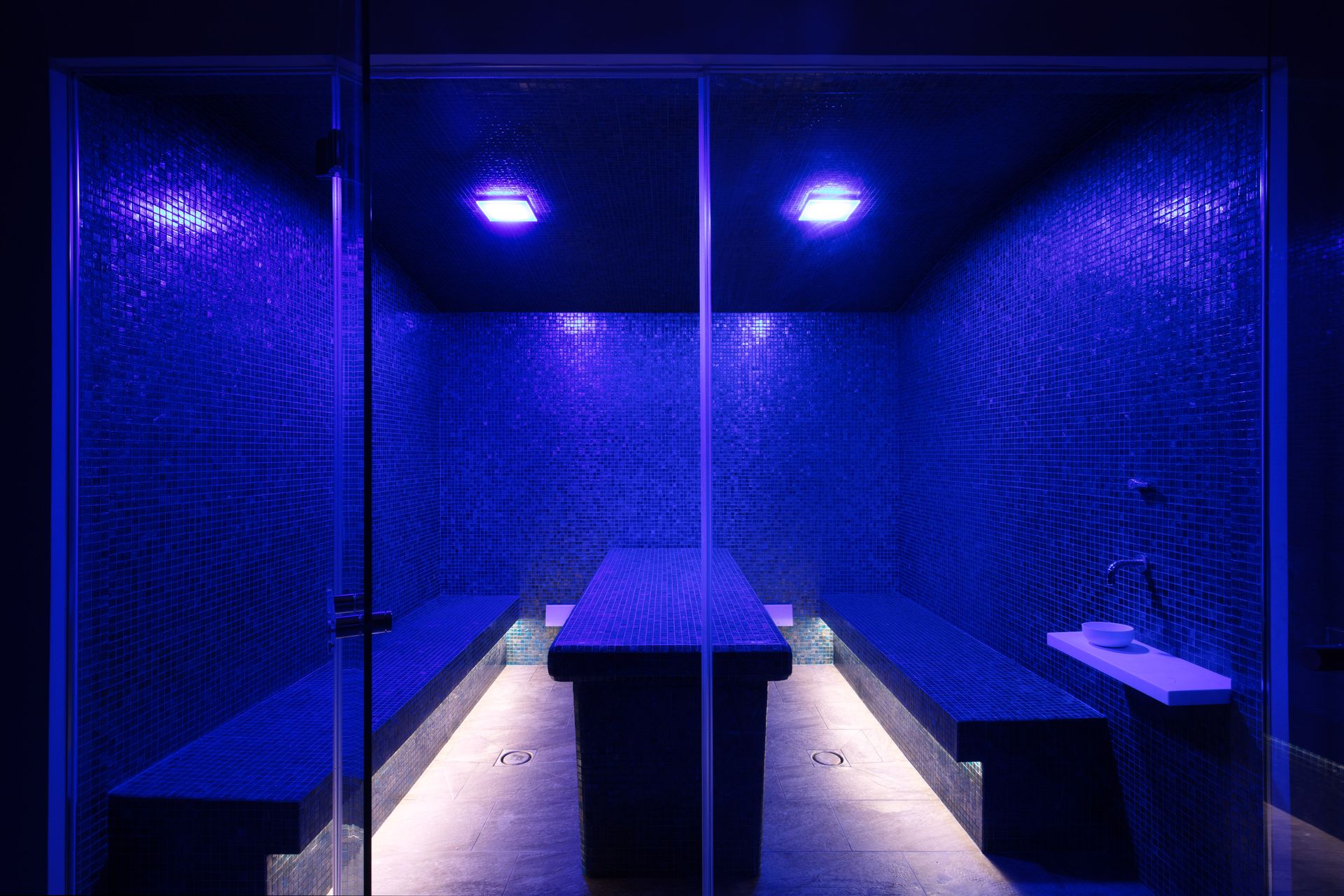 Una sauna a vapore con pareti rivestite di piastrelle blu, panche su entrambi i lati, un lettino per i trattamenti al centro e una soffusa illuminazione ambientale blu.