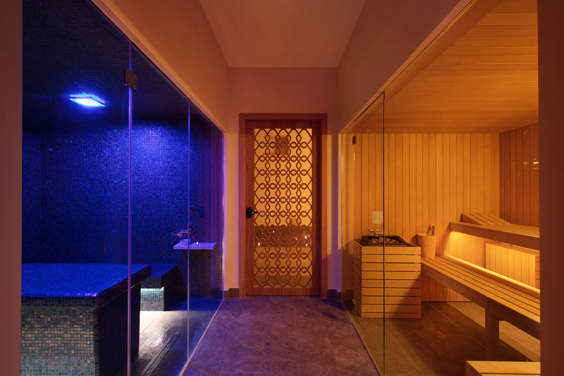 Un corridoio termale con un bagno turco illuminato di blu sulla sinistra, una sauna in legno sulla destra e al centro una porta a zanzariera decorativa.