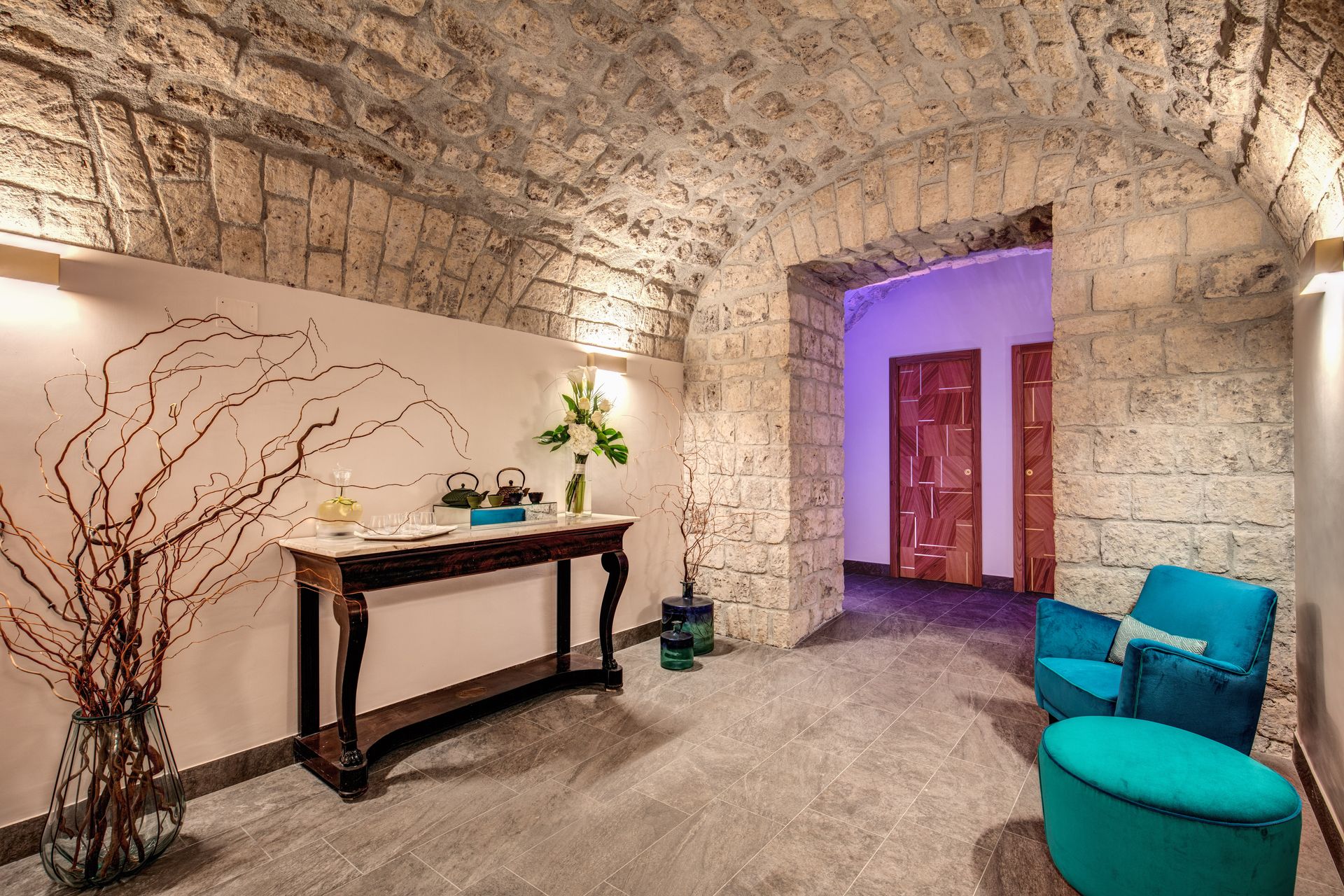 Un ingresso con volta in pietra, con una consolle in legno scuro, rami decorativi, una poltrona color verde acqua e un pouf.