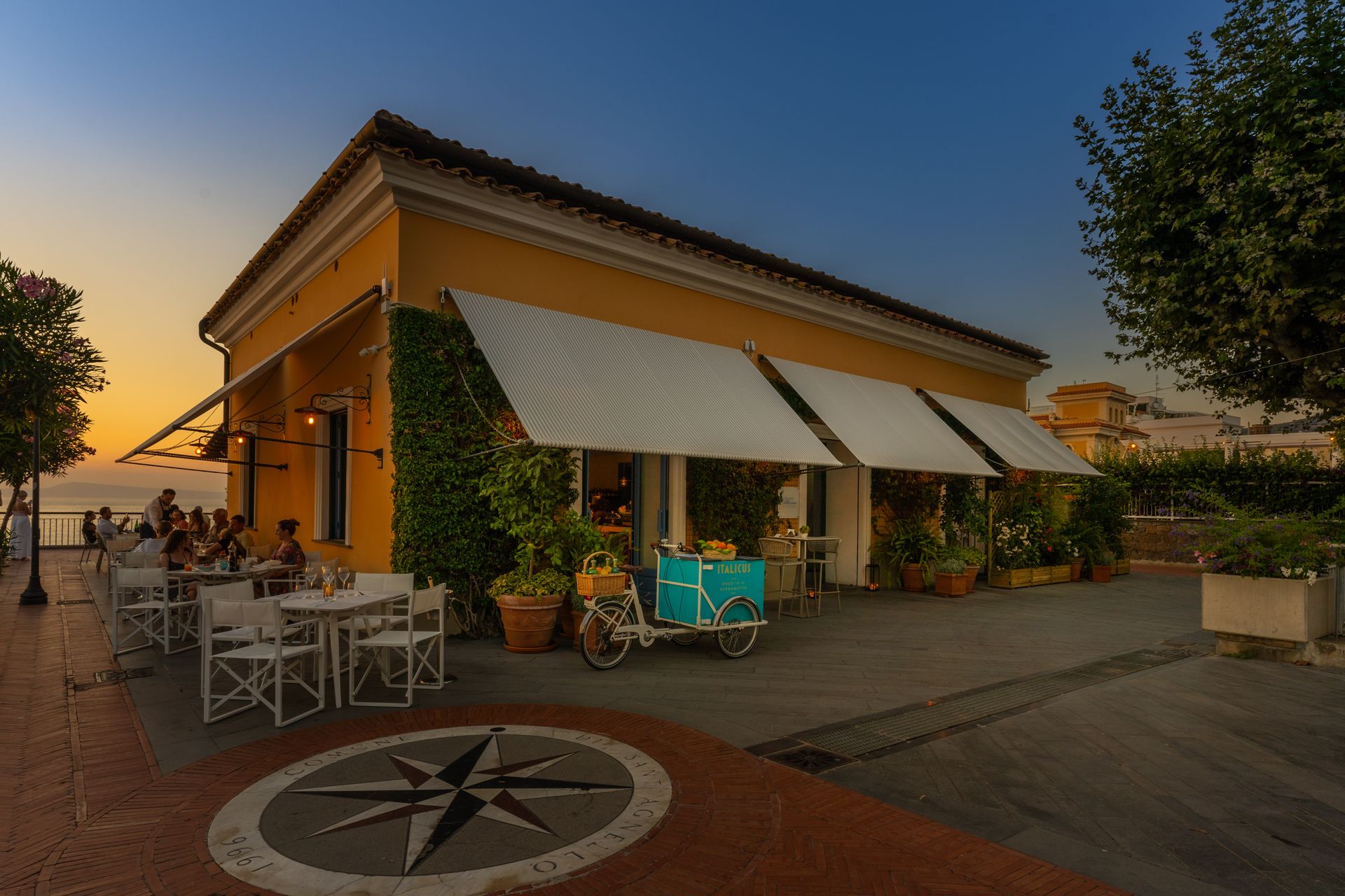 Vista al tramonto di un caffè con tavolini all'aperto, tende da sole a righe e piante in vaso su una terrazza.
