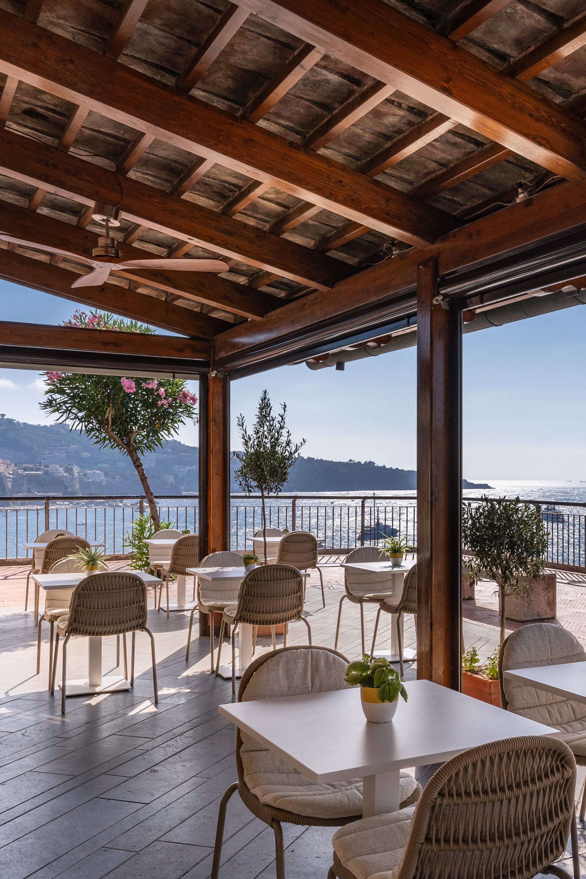 A sunny outdoor restaurant terrace with wicker chairs, white tables, and a wooden pergola overlooking the ocean.