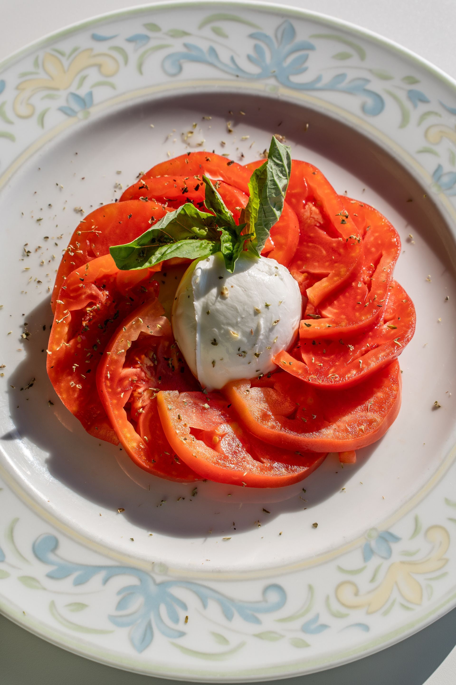 Sliced red tomatoes arranged in a circle on a patterned plate, topped with a ball of mozzarella and a basil sprig.