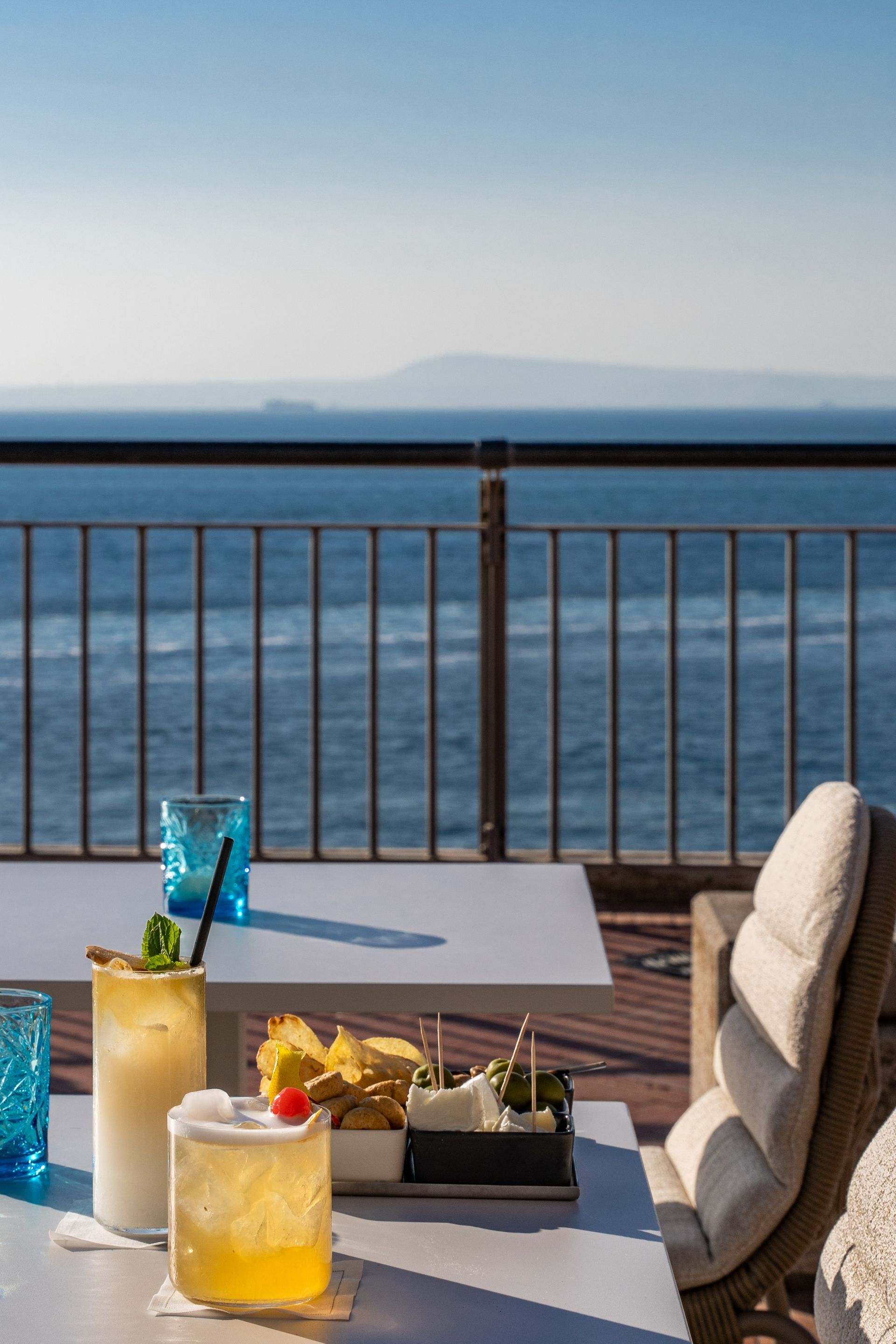 Two cocktails and a bowl of snacks on a patio table overlooking the ocean on a clear, sunny day.
