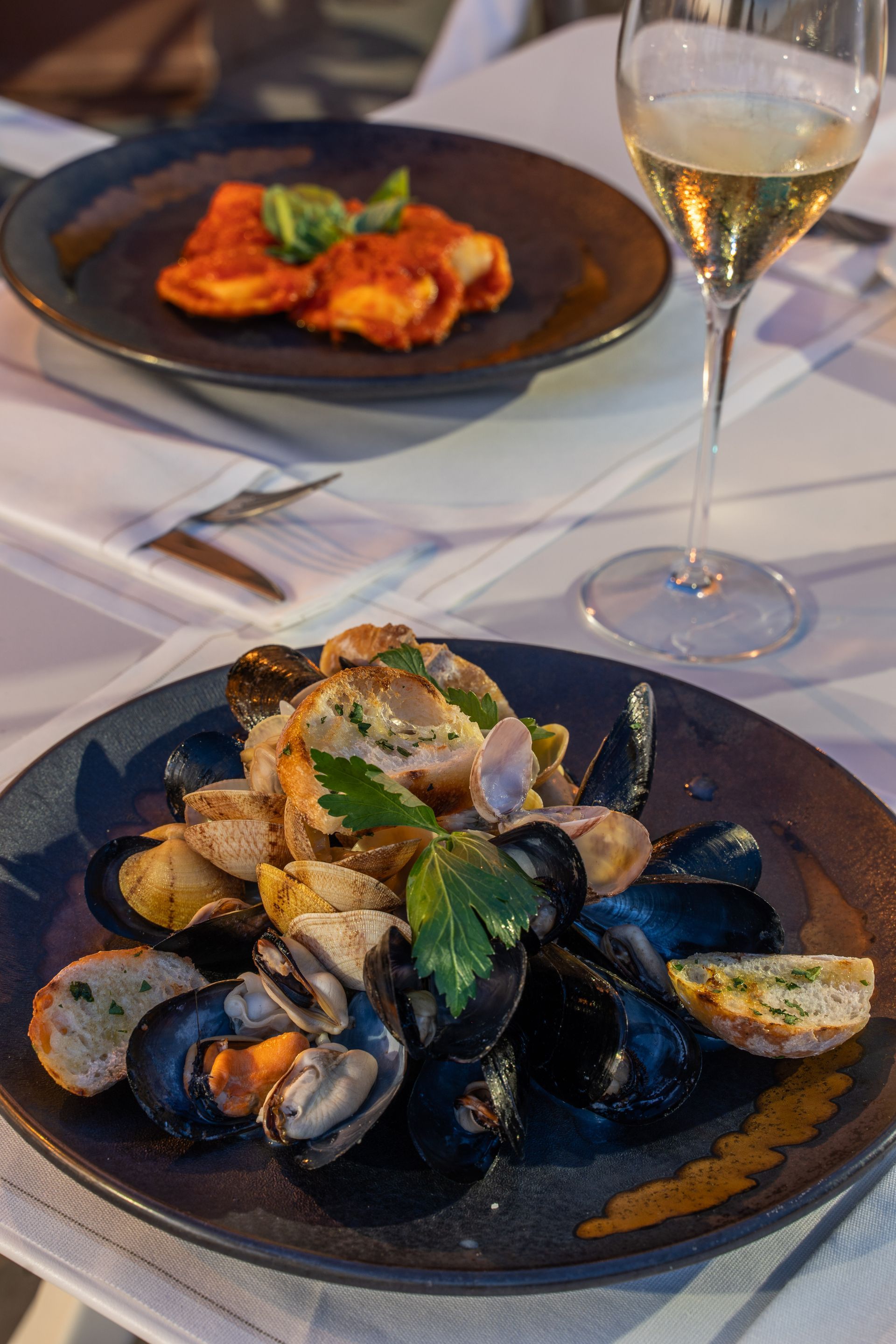 Due piatti gourmet su una tovaglia bianca, uno con cozze e pane, l'altro con pasta, accompagnati da un bicchiere di vino.
