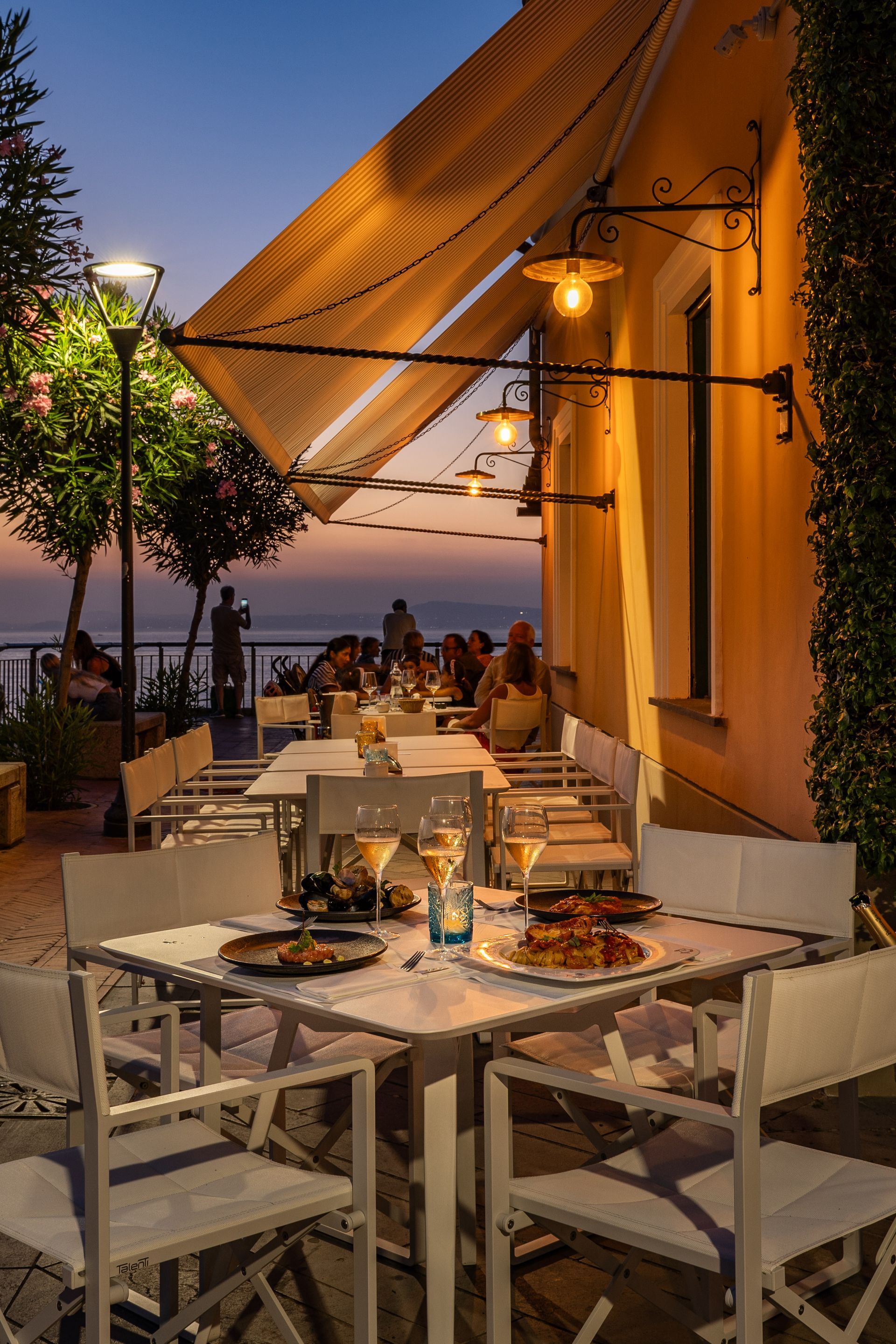 Tables set for dining on a sunset patio overlooking the sea, illuminated by warm wall-mounted lamps.
