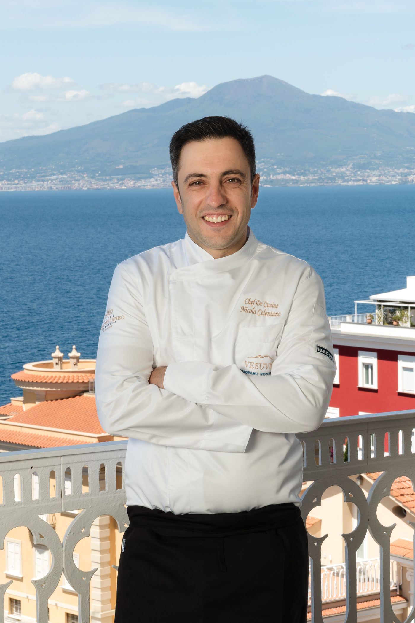 Uno chef sorridente in uniforme bianca è in piedi con le braccia incrociate su un balcone che si affaccia sul mare e su una montagna in lontananza.