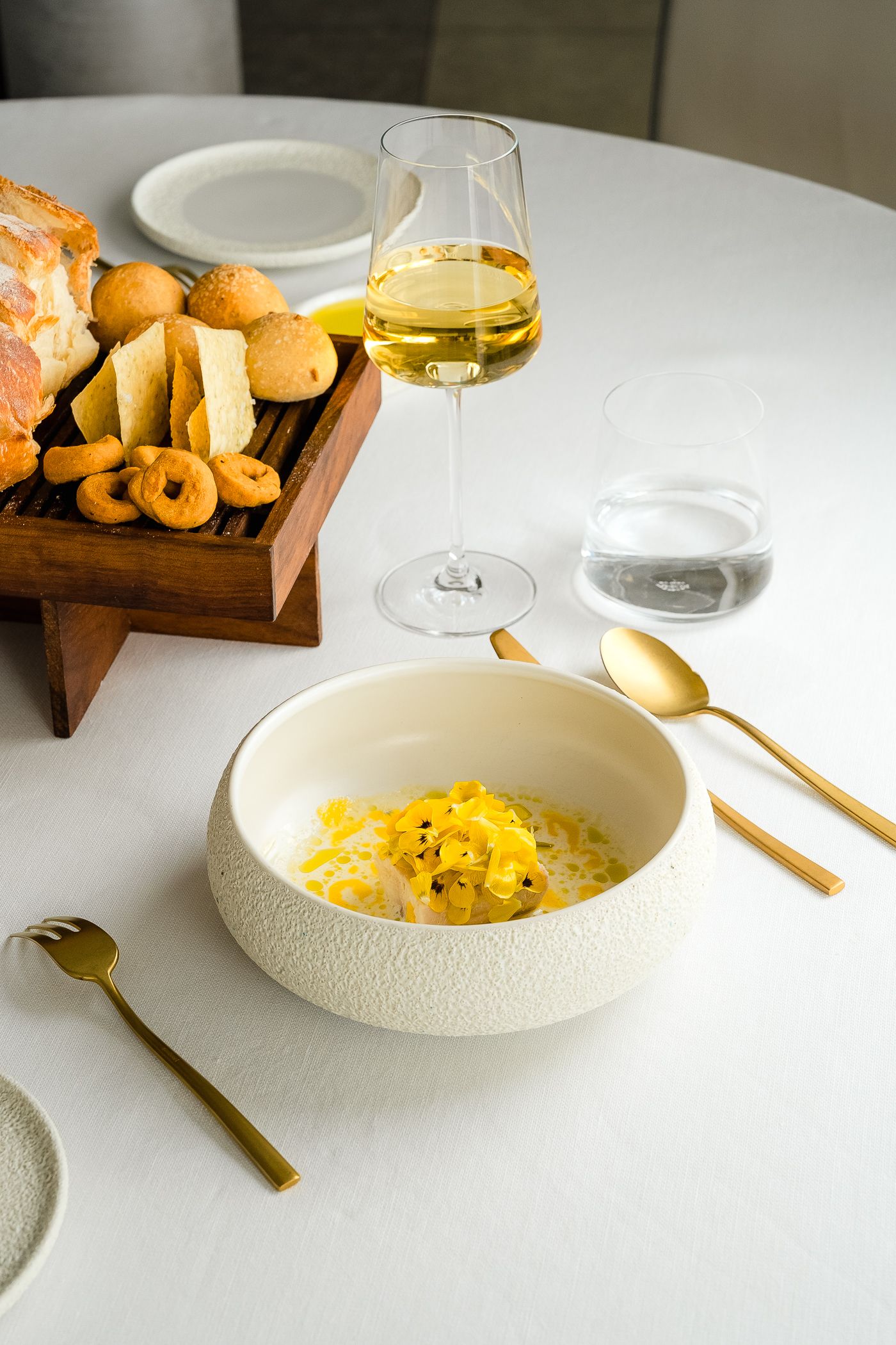 Una ciotola bianca con del risotto giallo è appoggiata su un tavolo accanto a un vassoio di legno con pane, vino e posate dorate.