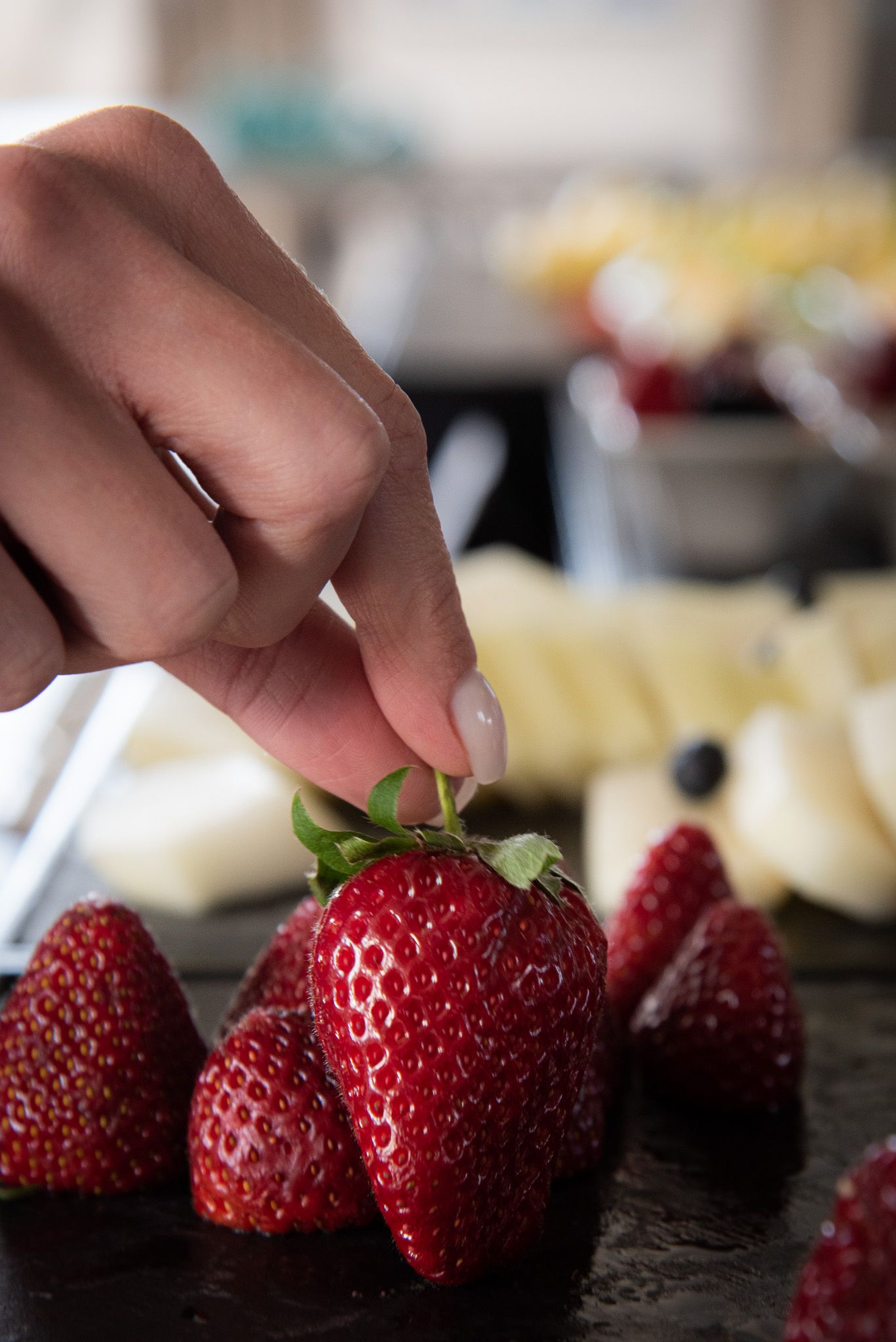 Una mano regge una fragola fresca e matura sopra un vassoio di frutta, tra cui fette di melone e mirtilli.