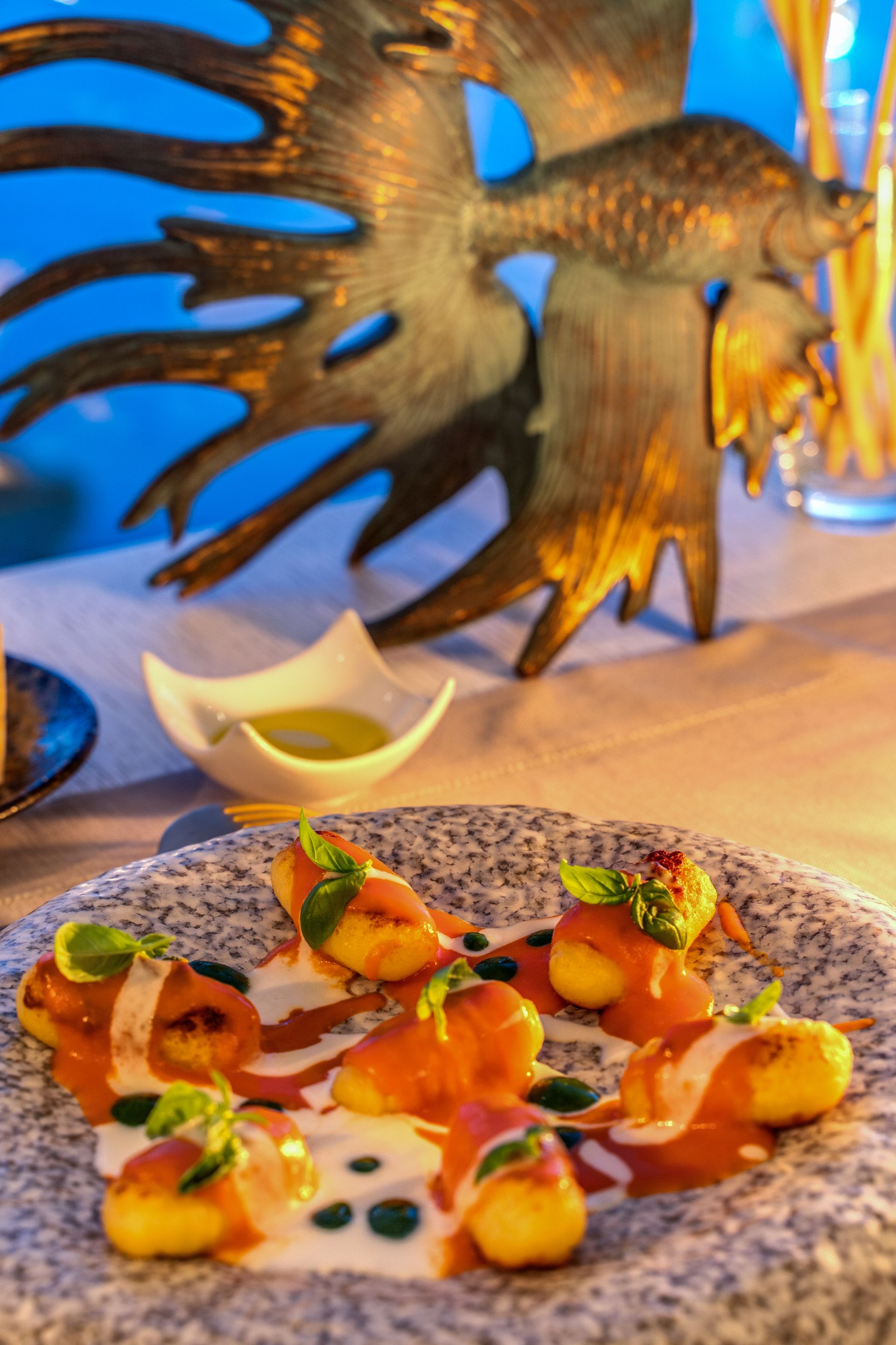 Gnocchi topped with tomato and cream sauces, garnished with basil on a textured gray plate near a bronze fish sculpture.