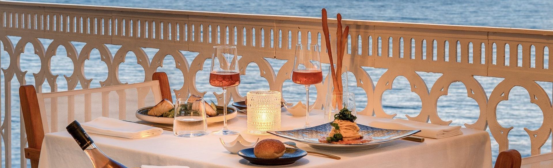 A table set for two with wine and food on a balcony overlooking the water.