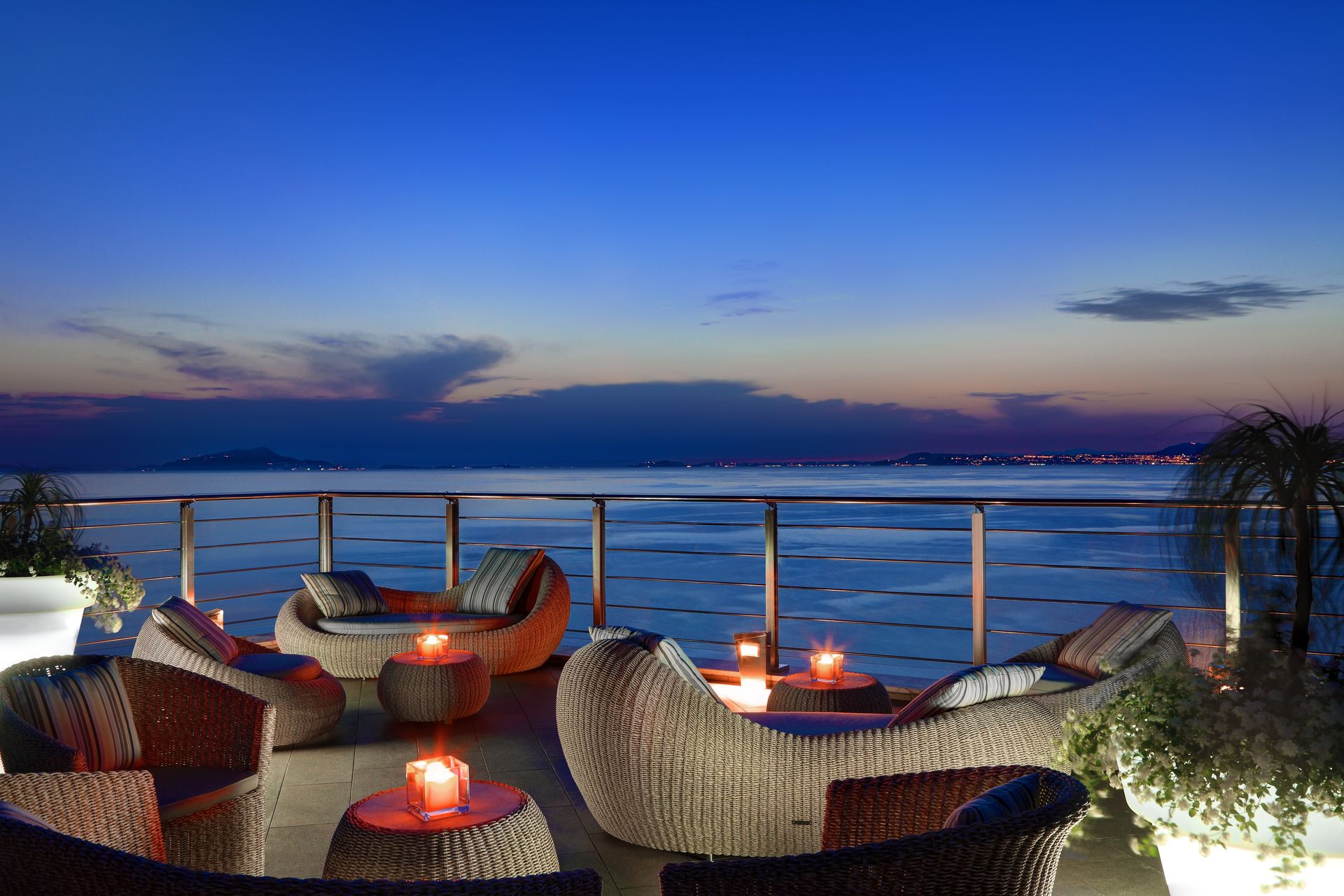 Set di mobili da salotto in vimini su una terrazza sul mare al crepuscolo, con lanterne luminose e un orizzonte calmo e di un blu intenso.